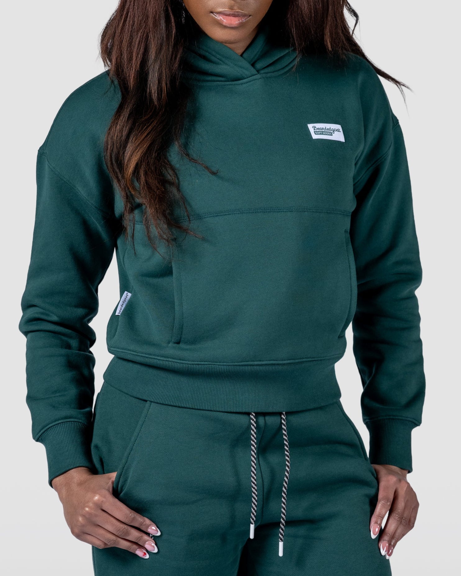 A woman with long dark hair wears the beardedgoat Womens Overnight Hoodie, standing against a plain light background with her hands in her pockets and looking down with a calm expression.