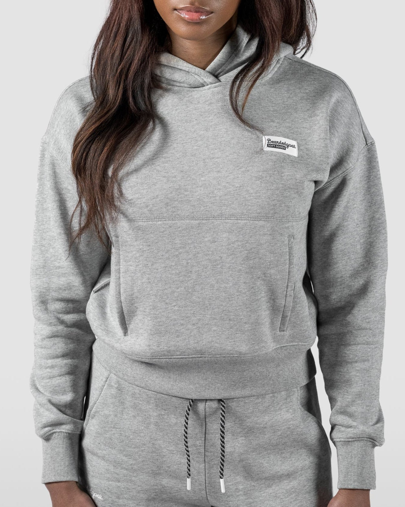 A woman wears the beardedgoat Women's Overnight™ Hoodie in light gray with a chest patch, paired with matching sweatpants, standing against a plain light background.
