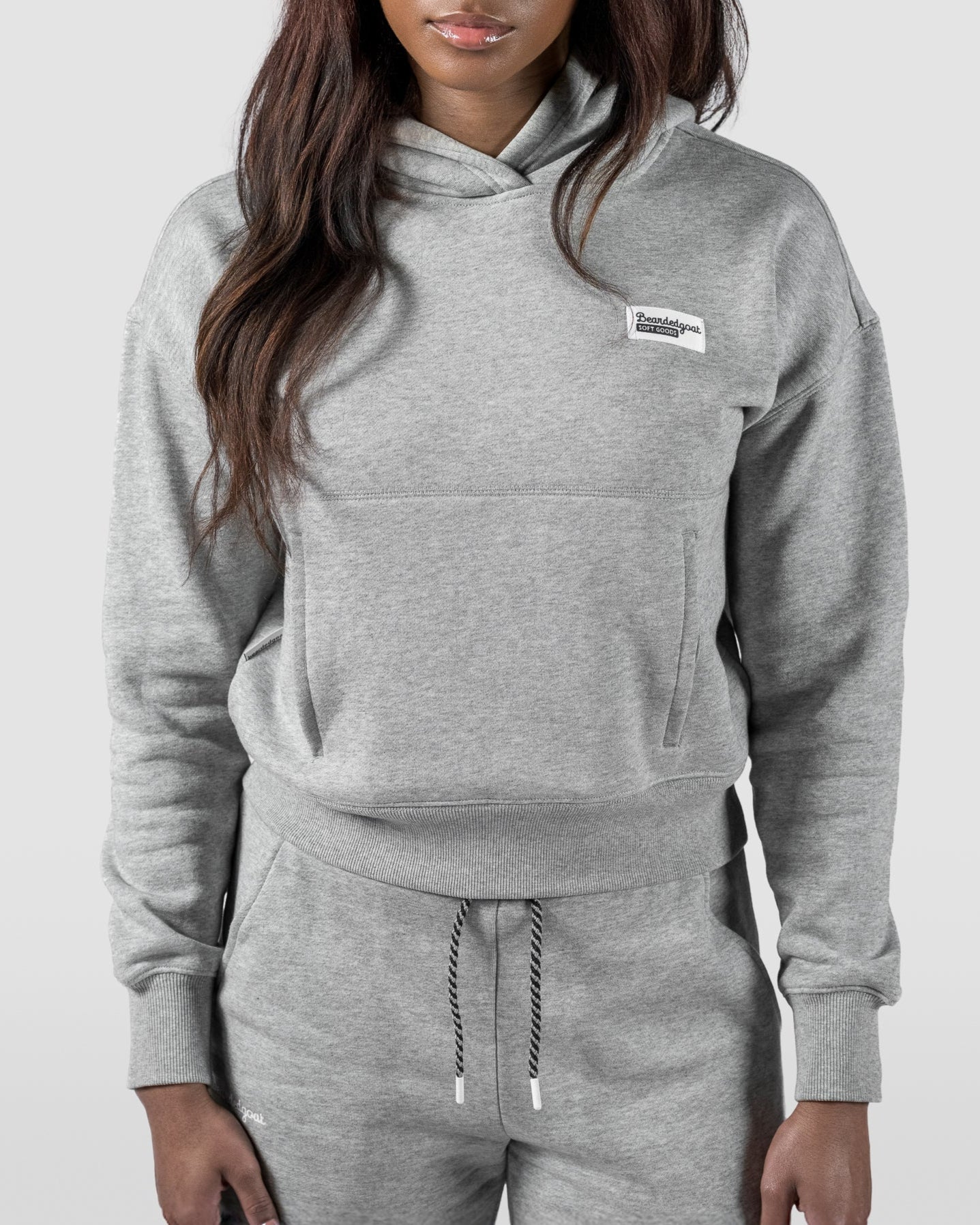 A woman with long dark hair wears the beardedgoat Womens Overnight Hoodie in light gray heavyweight cotton fleece, standing against a plain light background and facing the camera with a neutral expression.