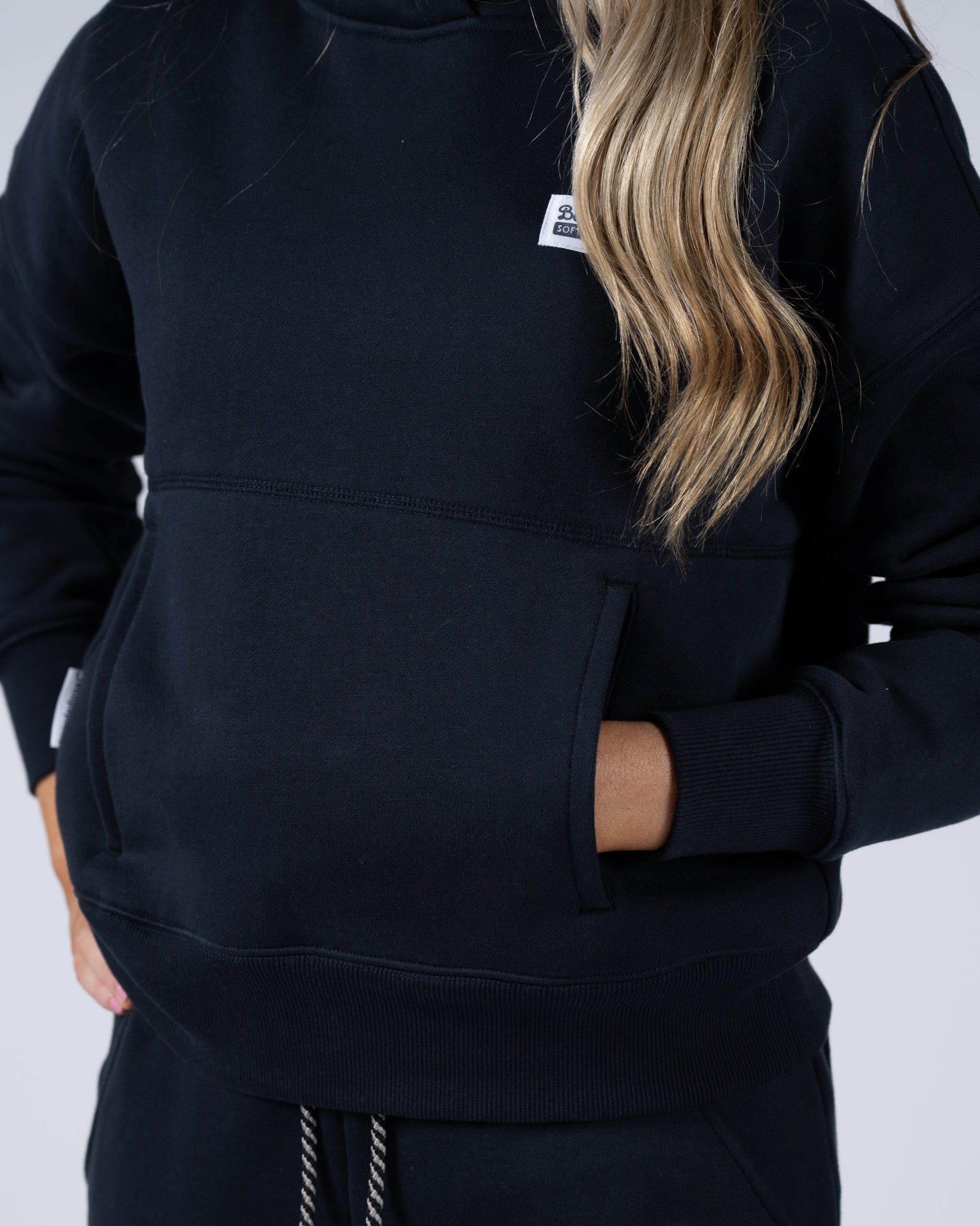 A BG woman with long, wavy blonde hair wears the beardedgoat Womens Overnight Hoodie, made of heavyweight cotton fleece, and matching joggers. Her hands are tucked in the front pocket. The plain background is light-colored.