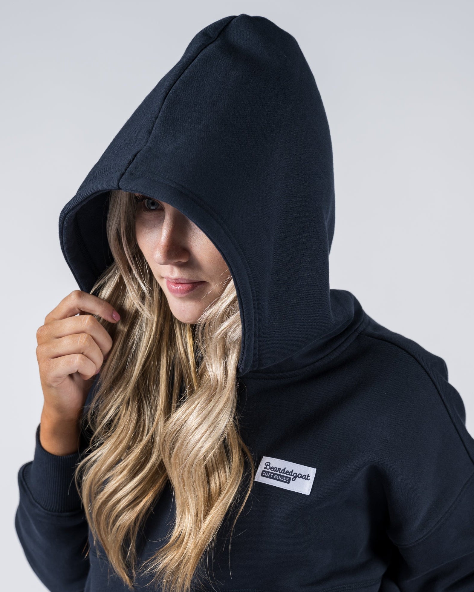 A BG woman with long, wavy blonde hair wears the beardedgoat Womens Overnight Hoodie in black heavyweight cotton fleece. The hood is up, partially covering her face, and a rectangular white label is on the chest. The background is plain light gray.