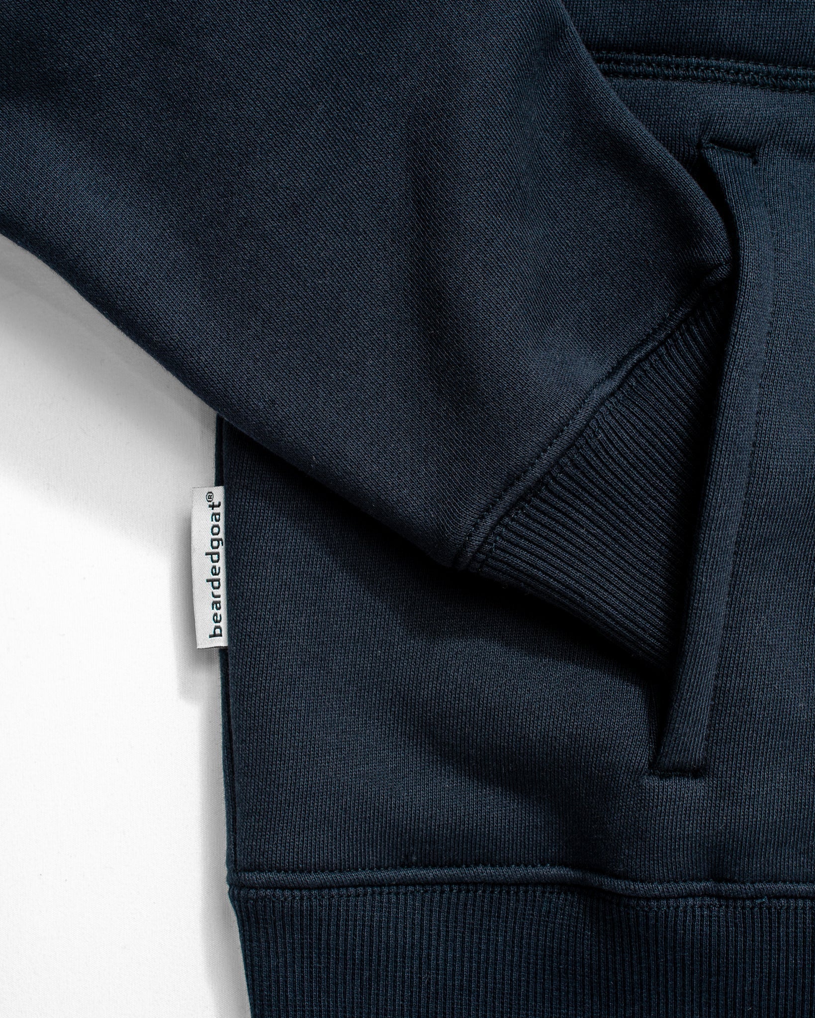 Close-up of the Womens Overnight Hoodie by beardedgoat in navy blue heavyweight cotton fleece, showing the pocket’s visible stitching, ribbed hem, and a small white beardedgoat tag sewn into the side seam near the pocket.