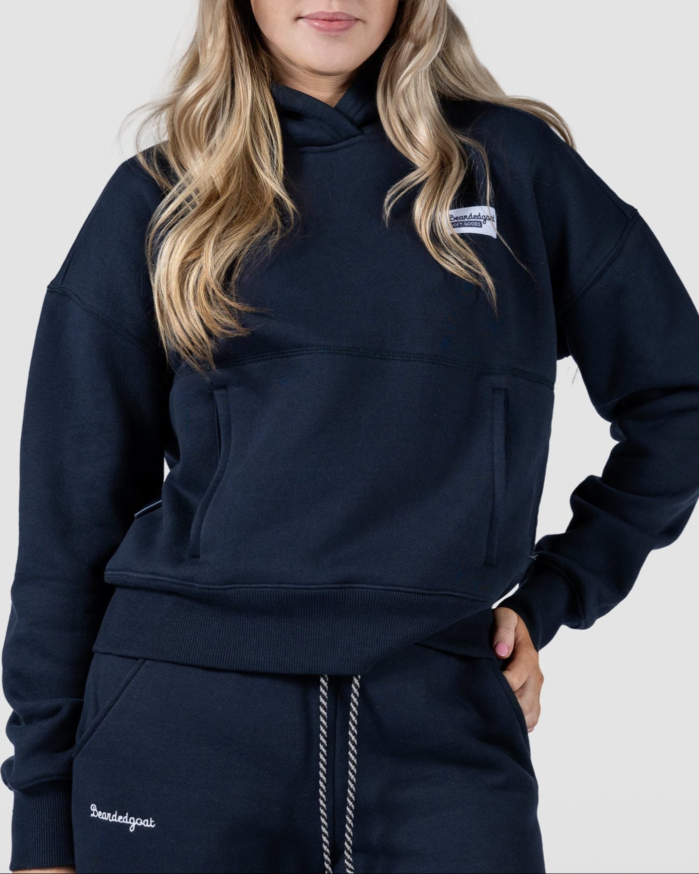 A woman with long blonde hair wears the beardedgoat Women's Overnight™ Hoodie and matching navy blue sweatpants, standing against a light gray background with one hand in her pocket, looking confidently at the camera.
