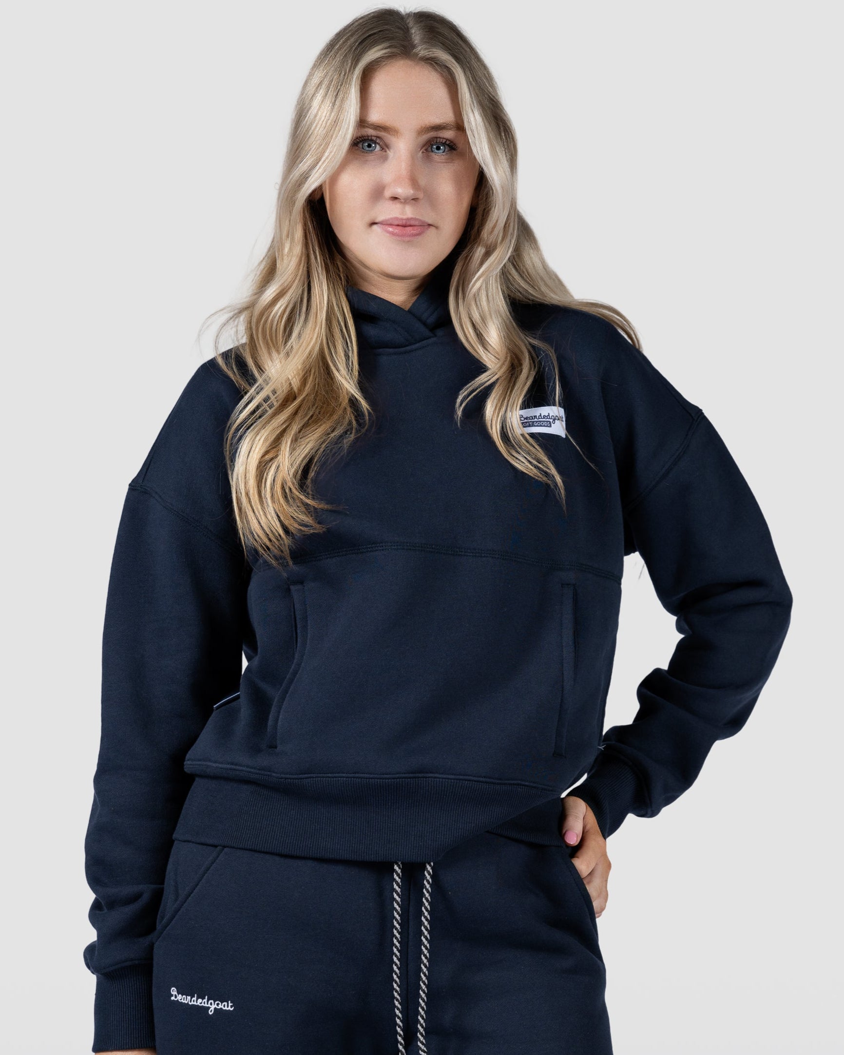 A BG woman with long blonde hair stands confidently against a plain light background, wearing the beardedgoat Womens Overnight Hoodie in dark navy heavyweight cotton fleece with matching sweatpants.