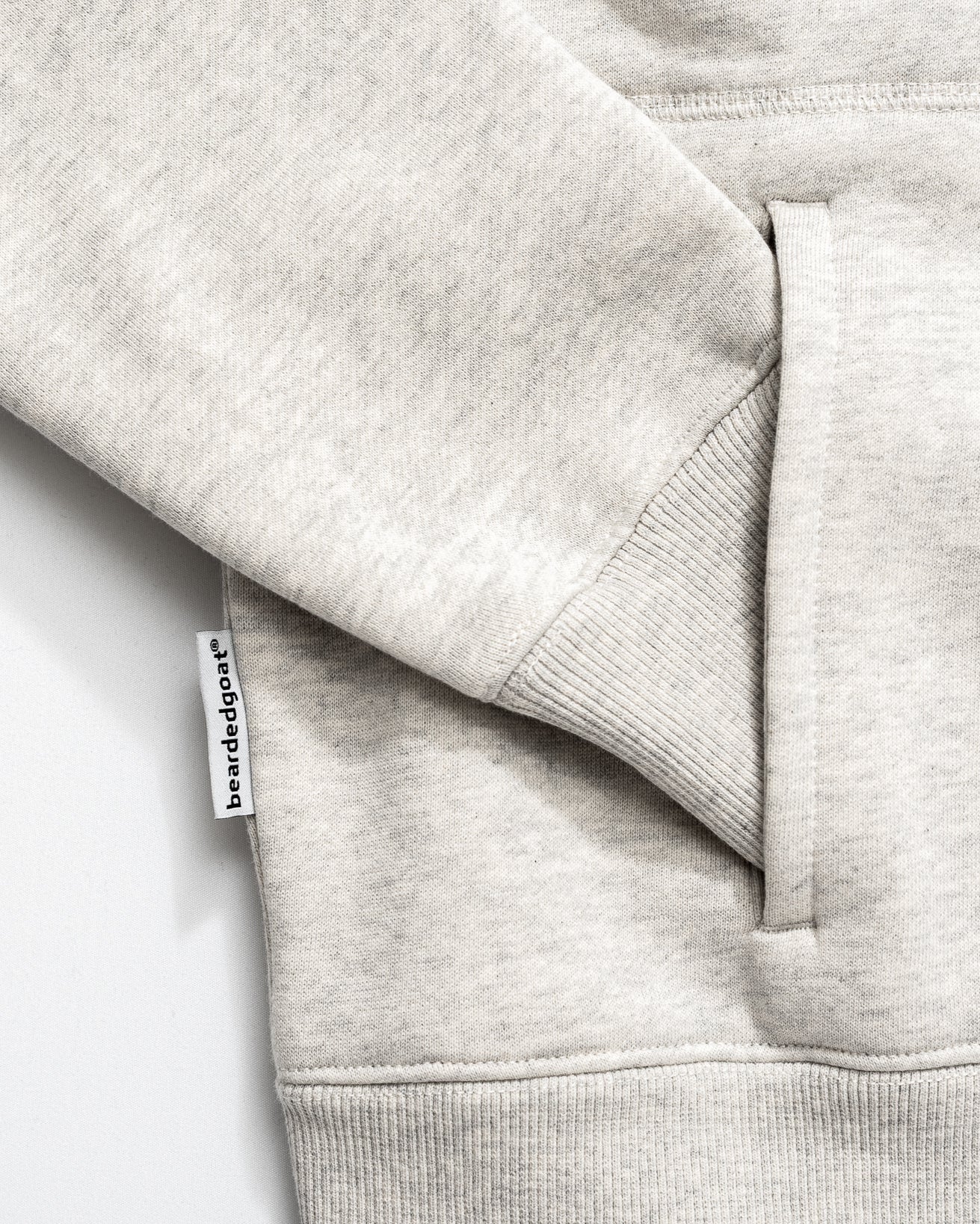 Close-up of the Womens Overnight Hoodie by beardedgoat in light gray heavyweight cotton fleece, highlighting the side pocket, visible seam, and a small white fabric tag reading “bareddoage”.