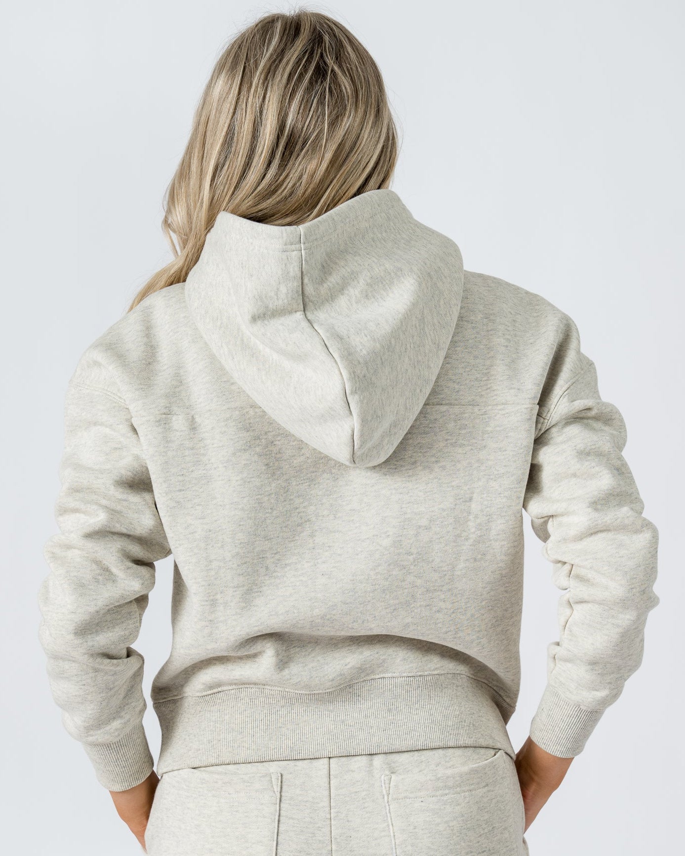 A woman with long blonde hair, seen from behind, wears the beardedgoat Womens Overnight Hoodie in light gray heavyweight cotton fleece against a plain white background.