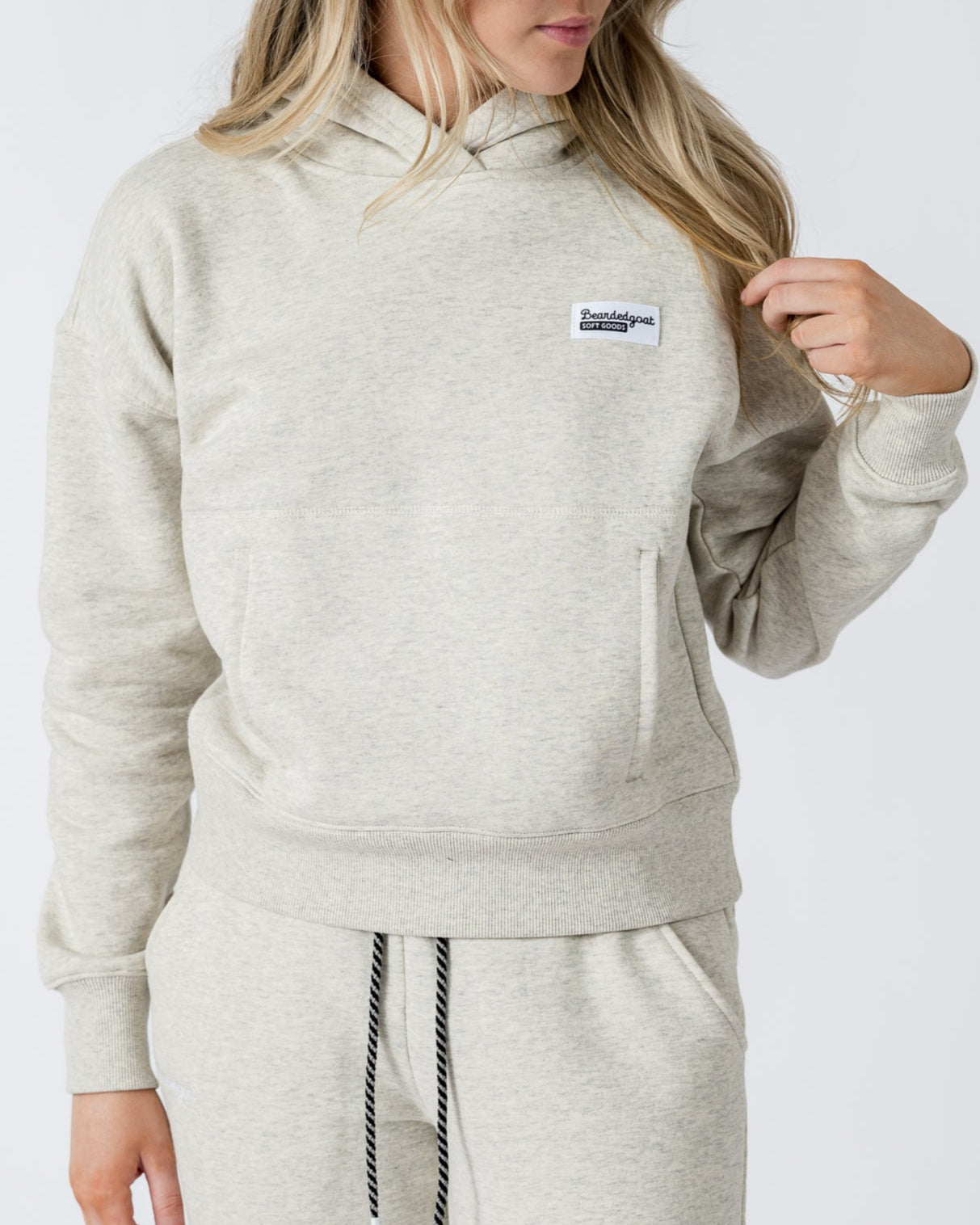 A woman with long blonde hair wears the beardedgoat Womens Overnight Hoodie in light gray heavyweight cotton fleece, featuring a small rectangular logo on the chest, paired with matching sweatpants against a plain background.