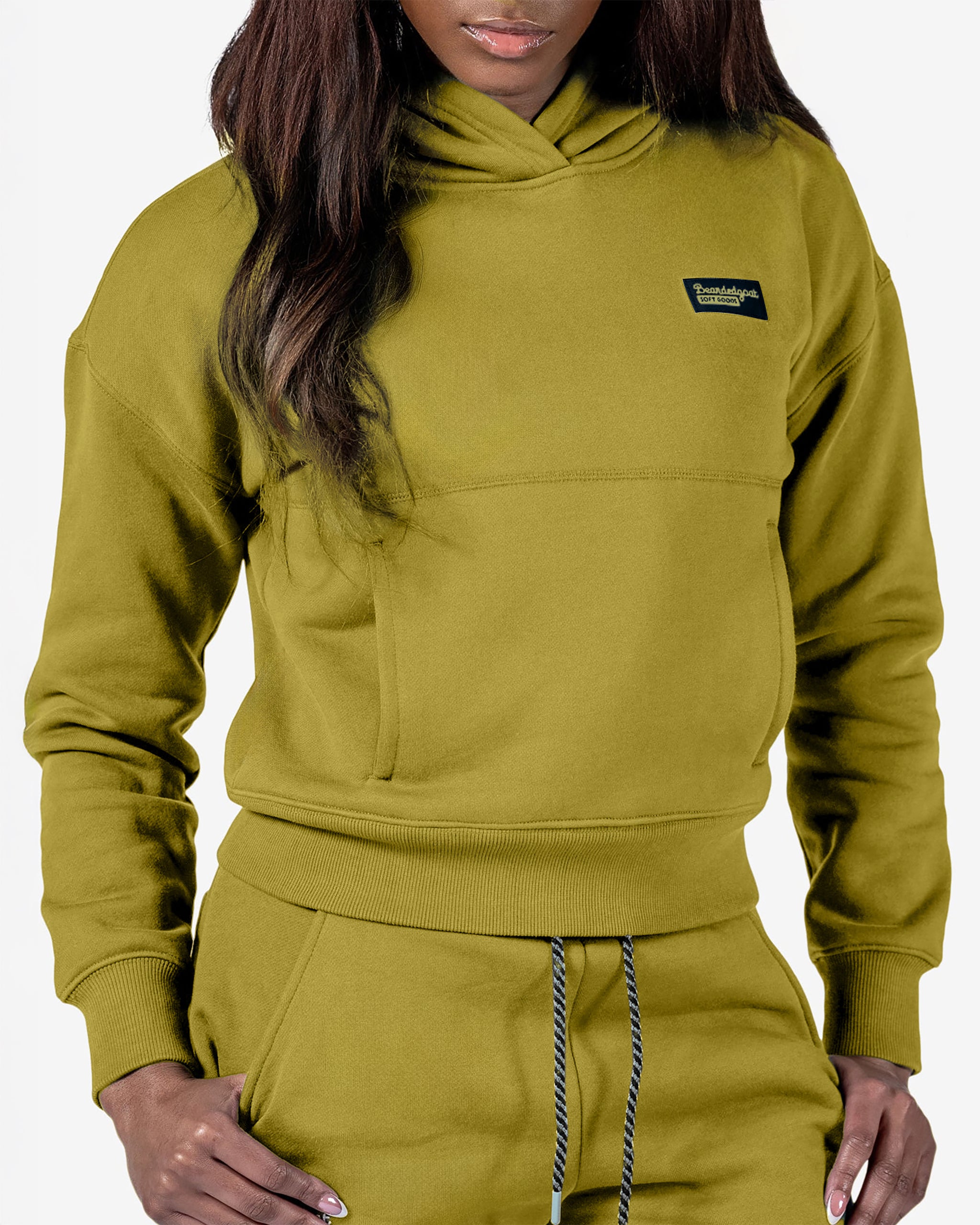 A person wears the beardedgoat Women's Overnight™ Hoodie in olive green with a black chest patch and matching sweatpants, both crafted from premium cotton fleece, standing against a plain white background.