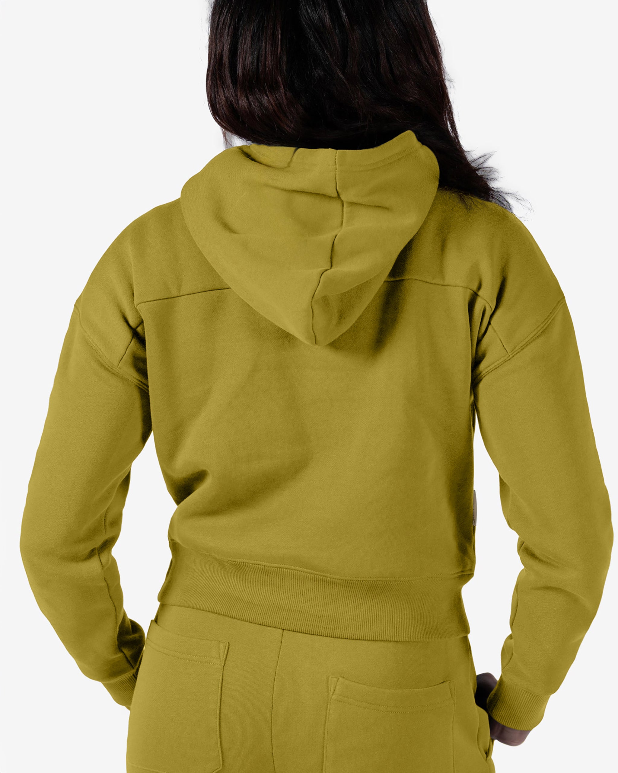 A woman with long dark hair faces away, wearing a relaxed fit olive green beardedgoat Women's Overnight™ Hoodie and sweatpants made from premium cotton fleece. The hood is down, hands on hips, against a plain white background.
