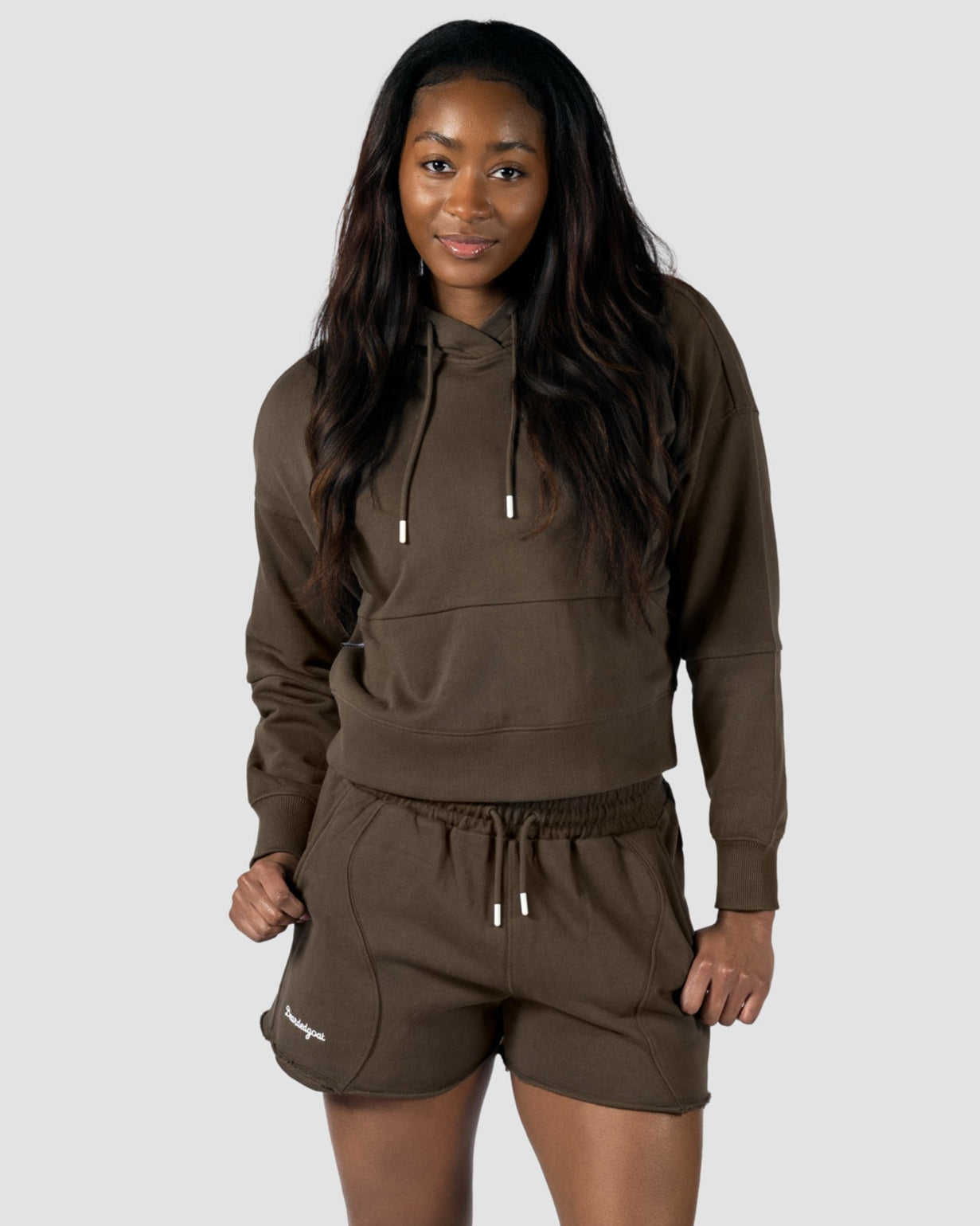 A woman faces forward in the Womens Drift Hoodie by BEARDEDGOAT, paired with matching shorts and white sneakers. The soft terry set, shown against a light gray backdrop, is perfect for spring-to-summer style.