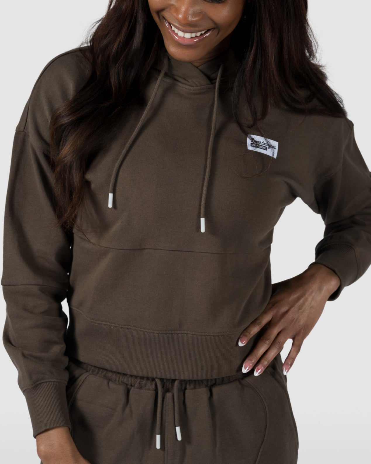 A woman smiles in the BEARDEDGOAT Womens Drift Hoodie, an olive green, lightweight terry set with a subtle chest patch—ideal for spring and summer. She stands against a plain light gray background.