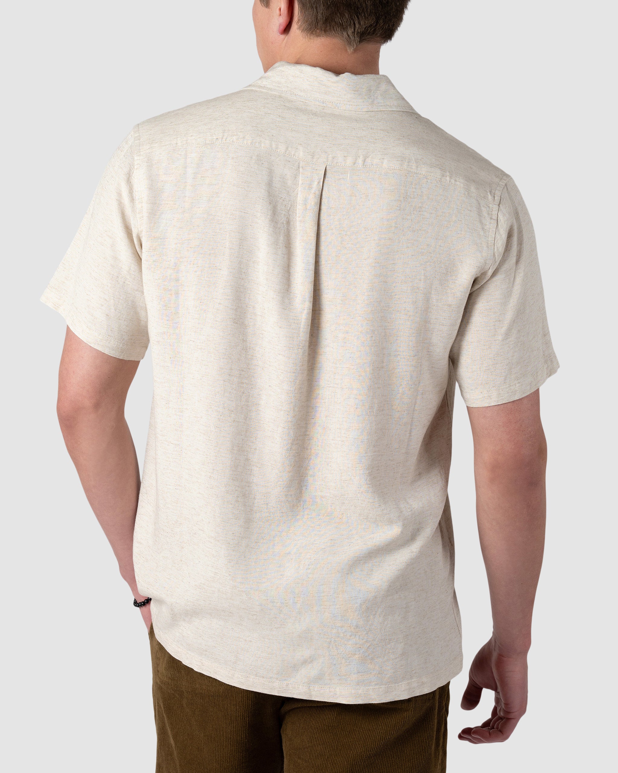 A man is shown from the back wearing the BEARDEDGOAT Mens Breeze Shirt, a lightweight, short-sleeve linen top in light beige with a subtle texture, straight hem, and upper back pleat. He pairs it with brown pants against a plain light gray background.