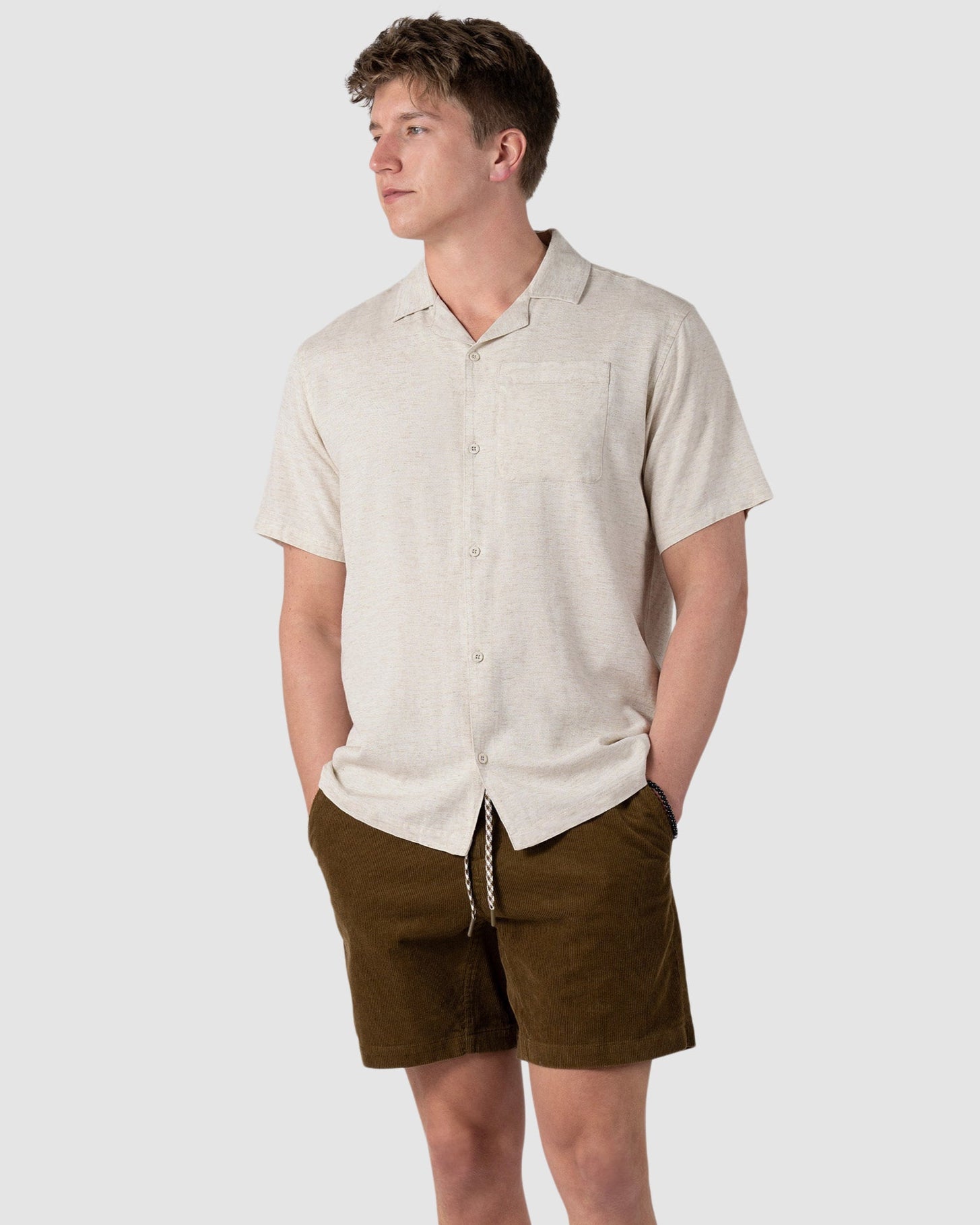 A young man stands with hands in pockets, wearing the BEARDEDGOAT Mens Breeze Shirt—a lightweight, short-sleeve beige button-up—paired with brown shorts and white sneakers against a plain light gray background.