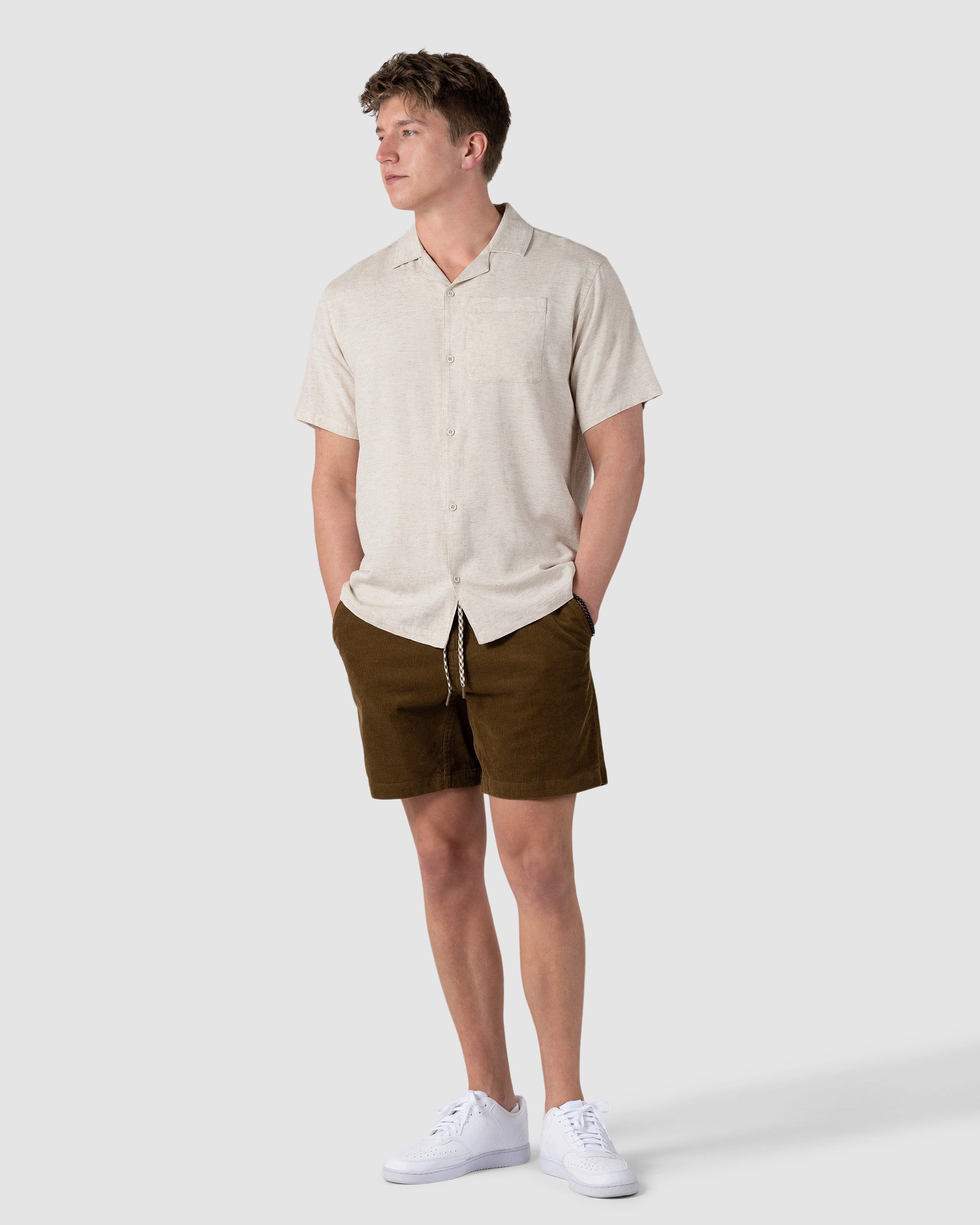 A young man stands with hands in pockets, wearing the BEARDEDGOAT Mens Breeze Shirt—a lightweight, short-sleeve beige button-up—paired with brown shorts and white sneakers against a plain light gray background.