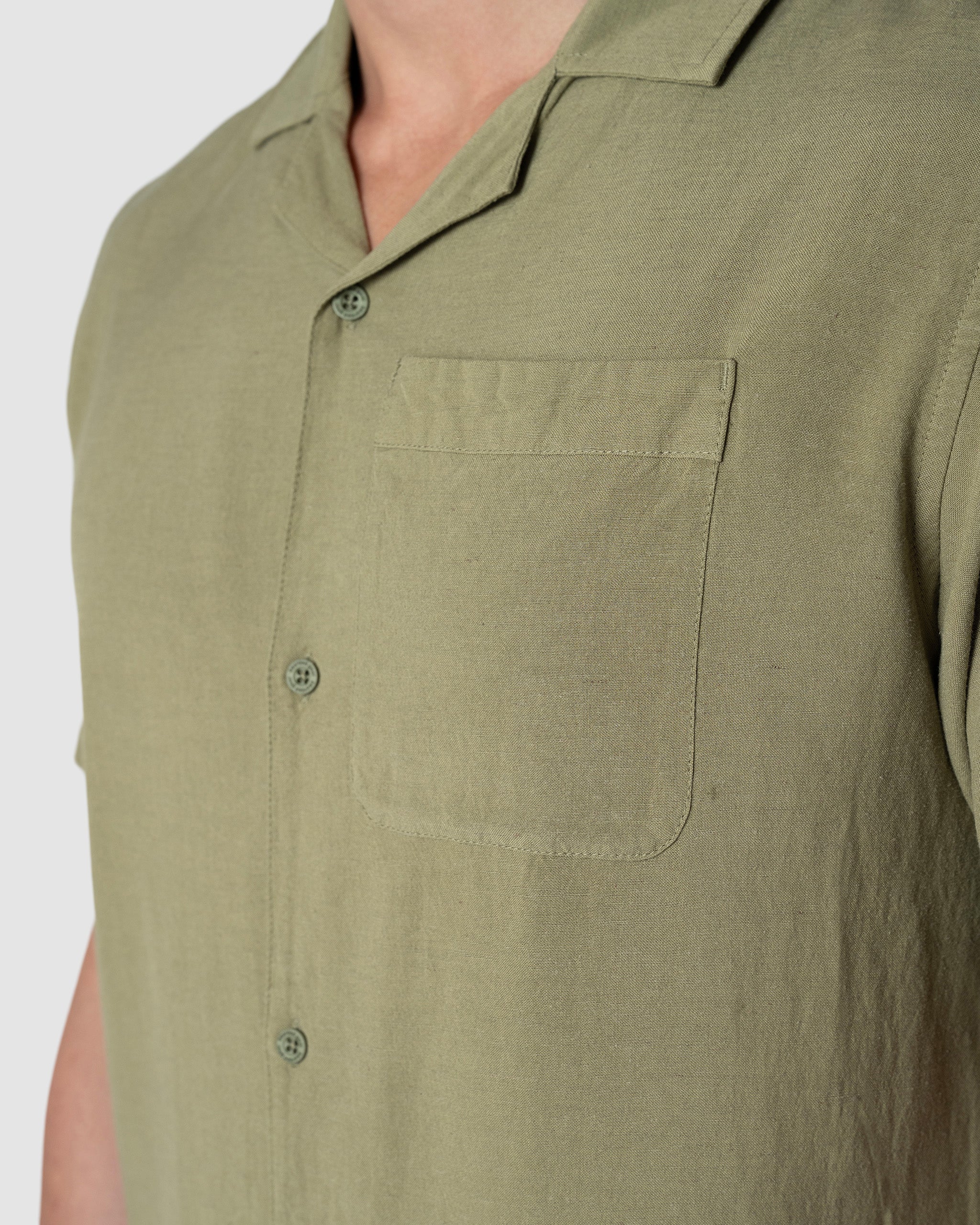 Close-up of a person wearing the BEARDEDGOAT Mens Breeze Shirt, a lightweight olive green short-sleeve button-up with chest pocket and collar, shown from shoulders to mid-torso against a plain light background.
