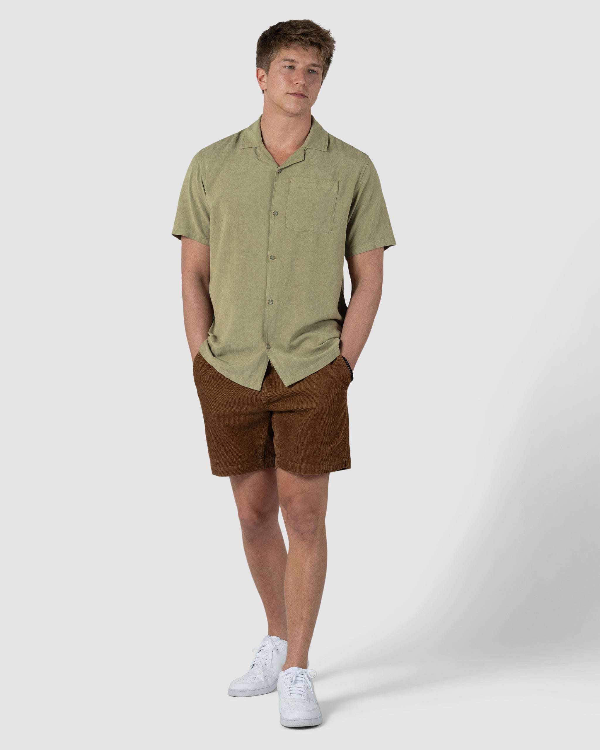 A young man stands with hands in pockets, wearing the BEARDEDGOAT Mens Breeze Shirt in light green linen, paired with brown shorts and white sneakers, against a plain light gray background.