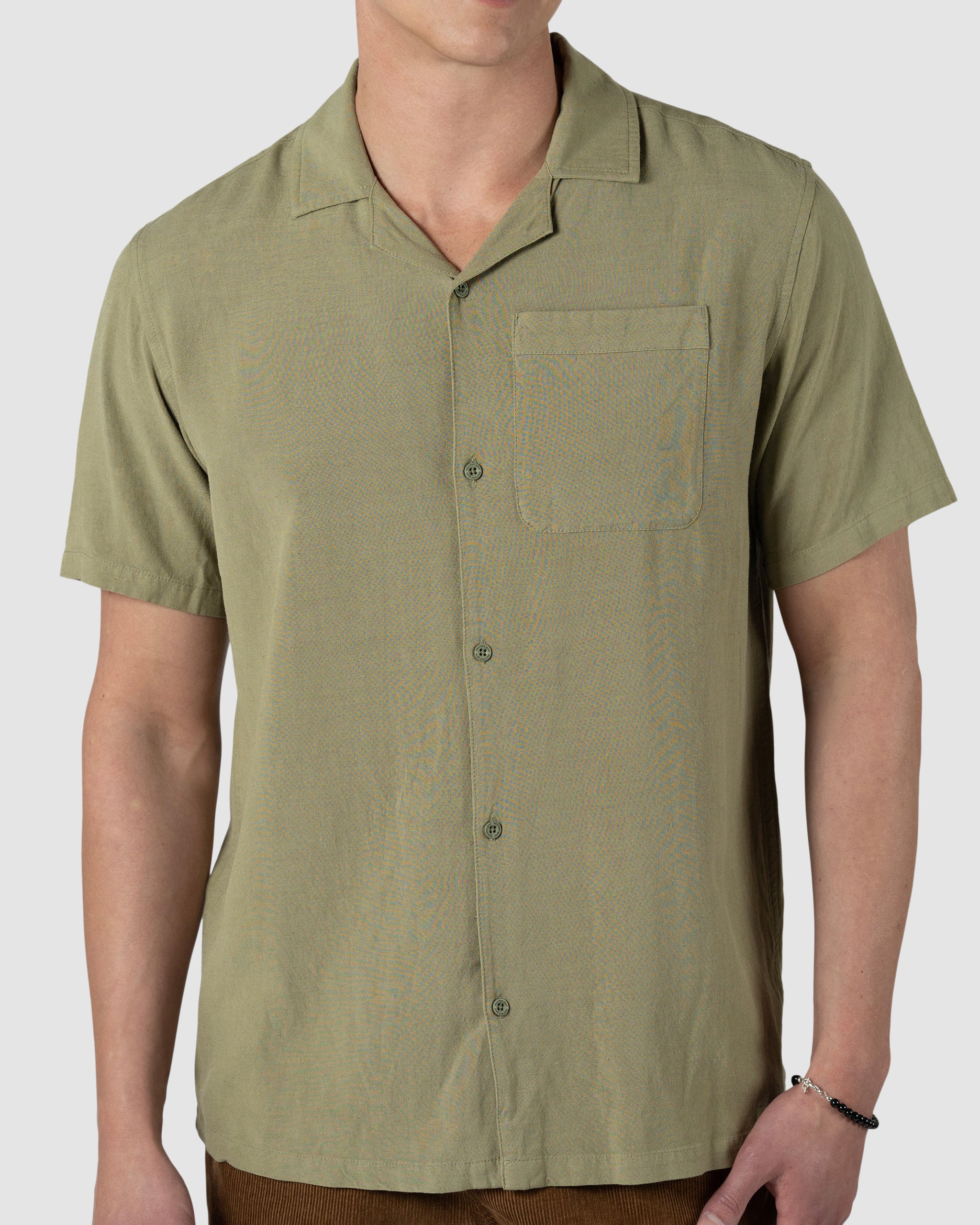 A person models the BEARDEDGOAT Mens Breeze Shirt, an olive green lightweight, breathable linen short-sleeve button-up with a chest pocket, standing against a plain light background. The persons head is not shown in the image.