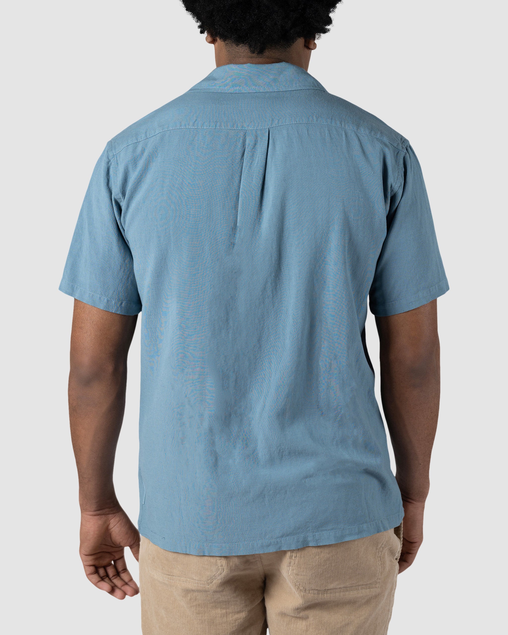 A person with short curly hair, seen from behind against a plain light gray background, wears the BEARDEDGOAT Mens Breeze Shirt in light blue and beige pants.