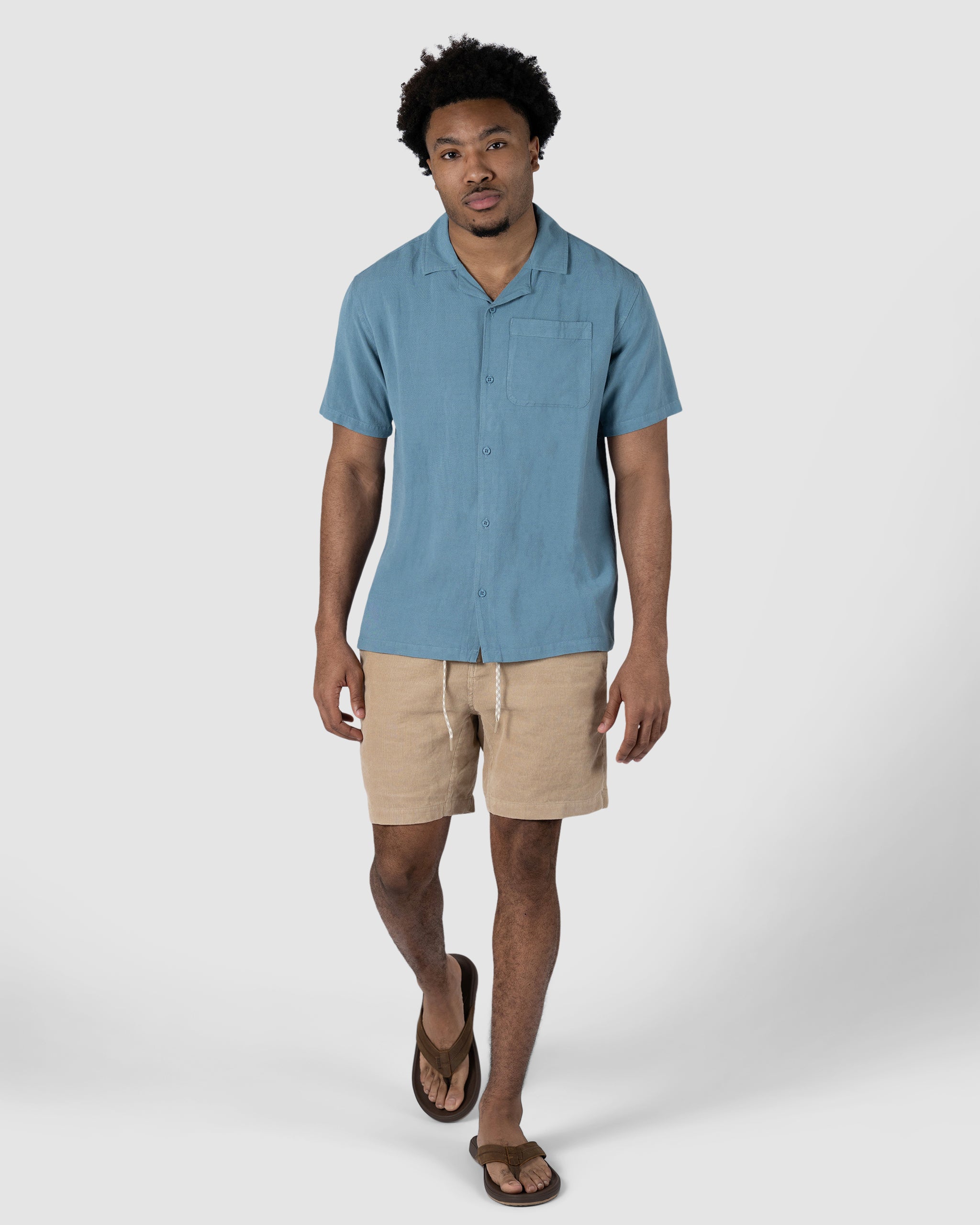 A man stands against a plain white background wearing the BEARDEDGOAT Mens Breeze Shirt, a lightweight light blue short-sleeve button-up, paired with breathable beige linen drawstring shorts and brown sandals.