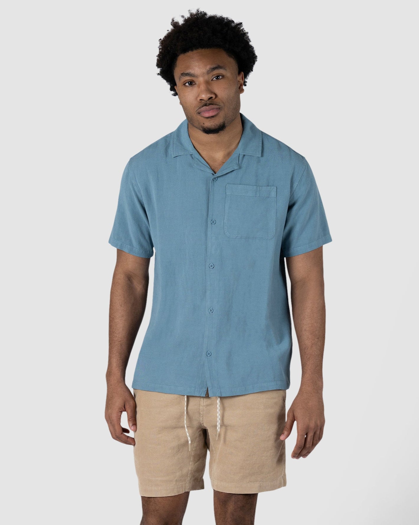 A man stands against a plain white background wearing the BEARDEDGOAT Mens Breeze Shirt, a lightweight light blue short-sleeve button-up, paired with breathable beige linen drawstring shorts and brown sandals.