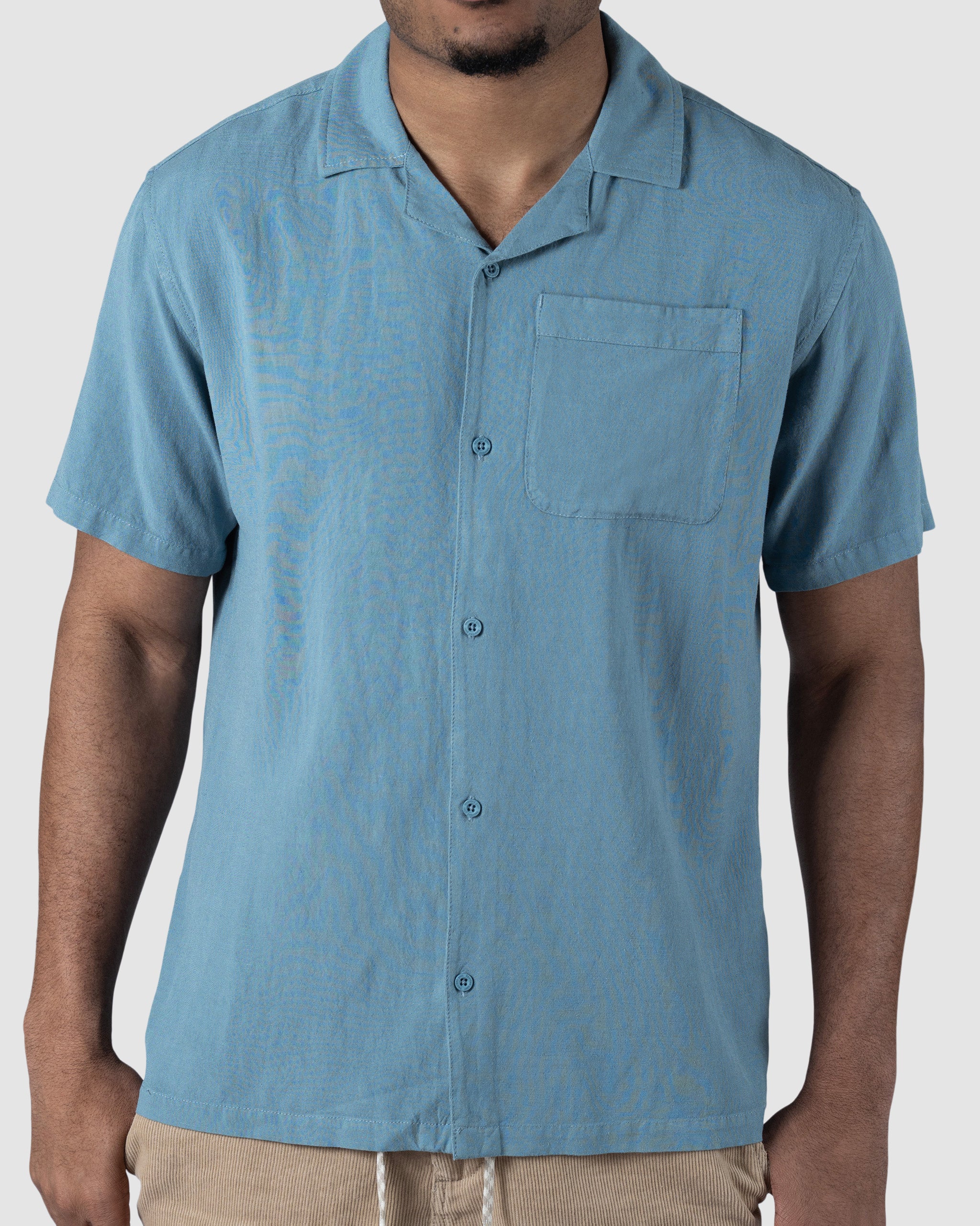 A man wears the BEARDEDGOAT Mens Breeze Shirt—a light blue, short-sleeved linen button-up with a chest pocket—against a plain background. The breathable, lightweight fabric ensures comfort; his face is partially out of frame.