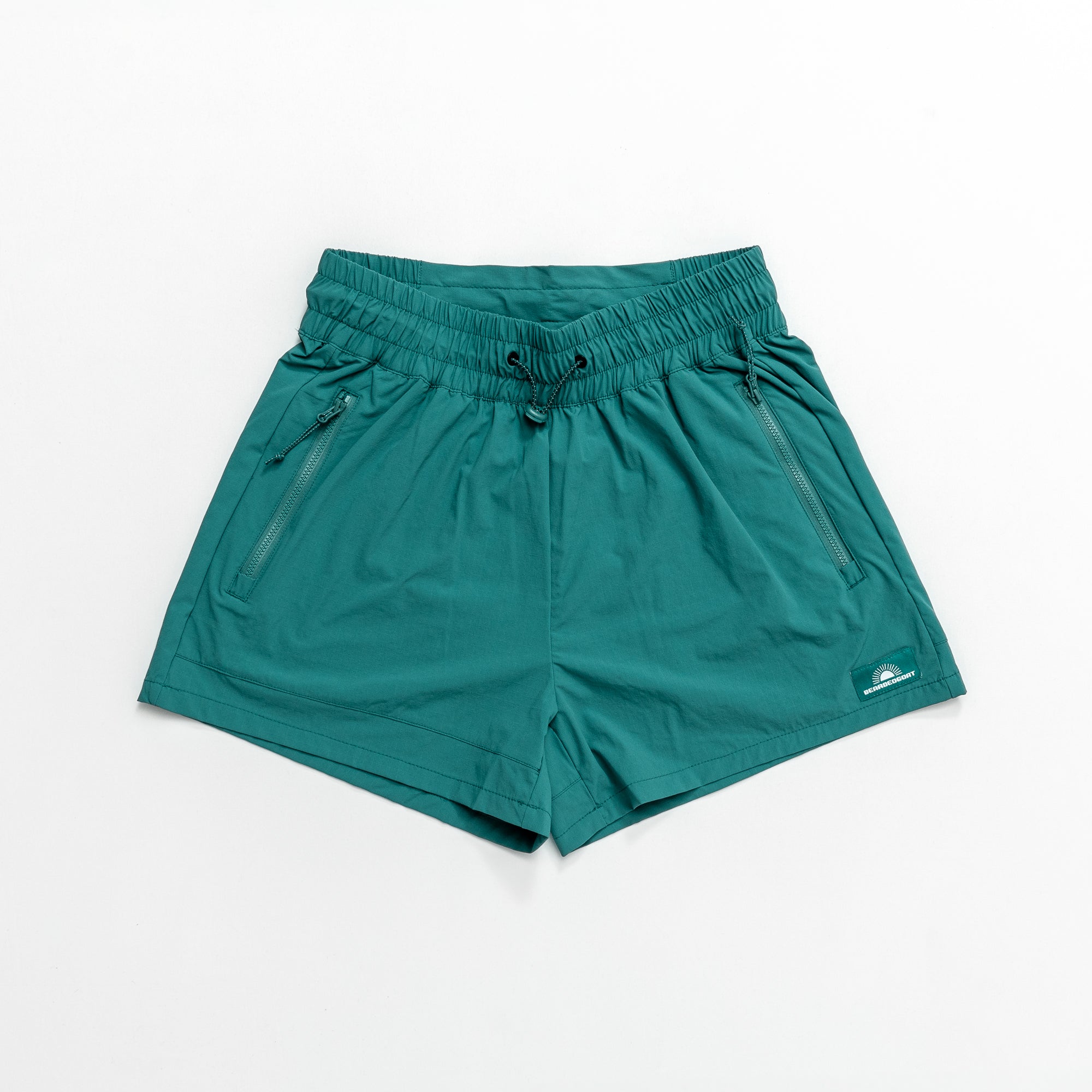 The beardedgoat Womens Alpina Short in teal green features a high-waisted fit, stretchy ripstop fabric, elastic waistband with drawstring, two zippered front pockets, and a small logo near the left hem. Displayed flat on a white background.