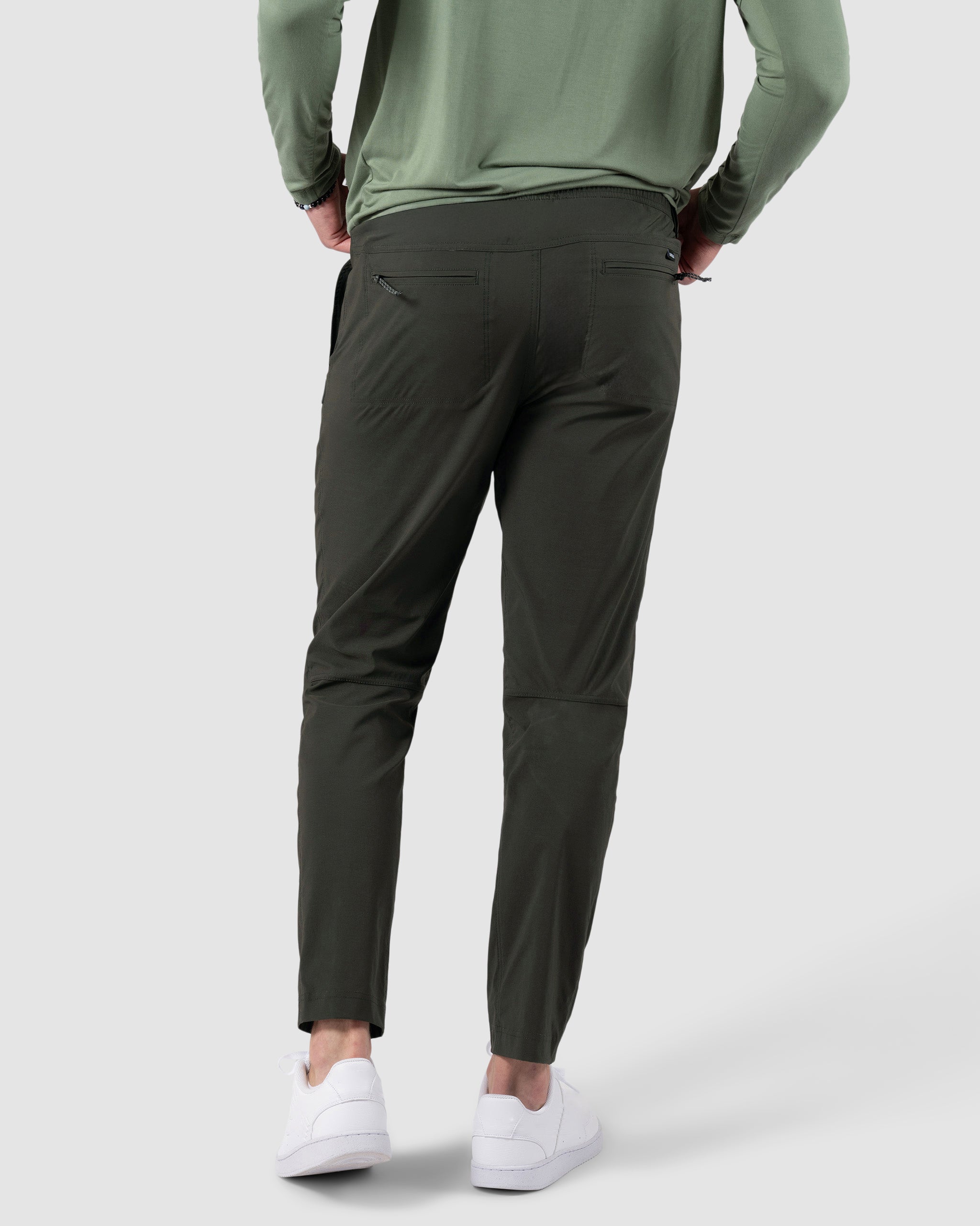 A person in a light green long-sleeve shirt, BEARDEDGOAT NEW-—ROVER™ Pant in dark green, and white sneakers stands facing away with hands on hips before a plain light gray background.