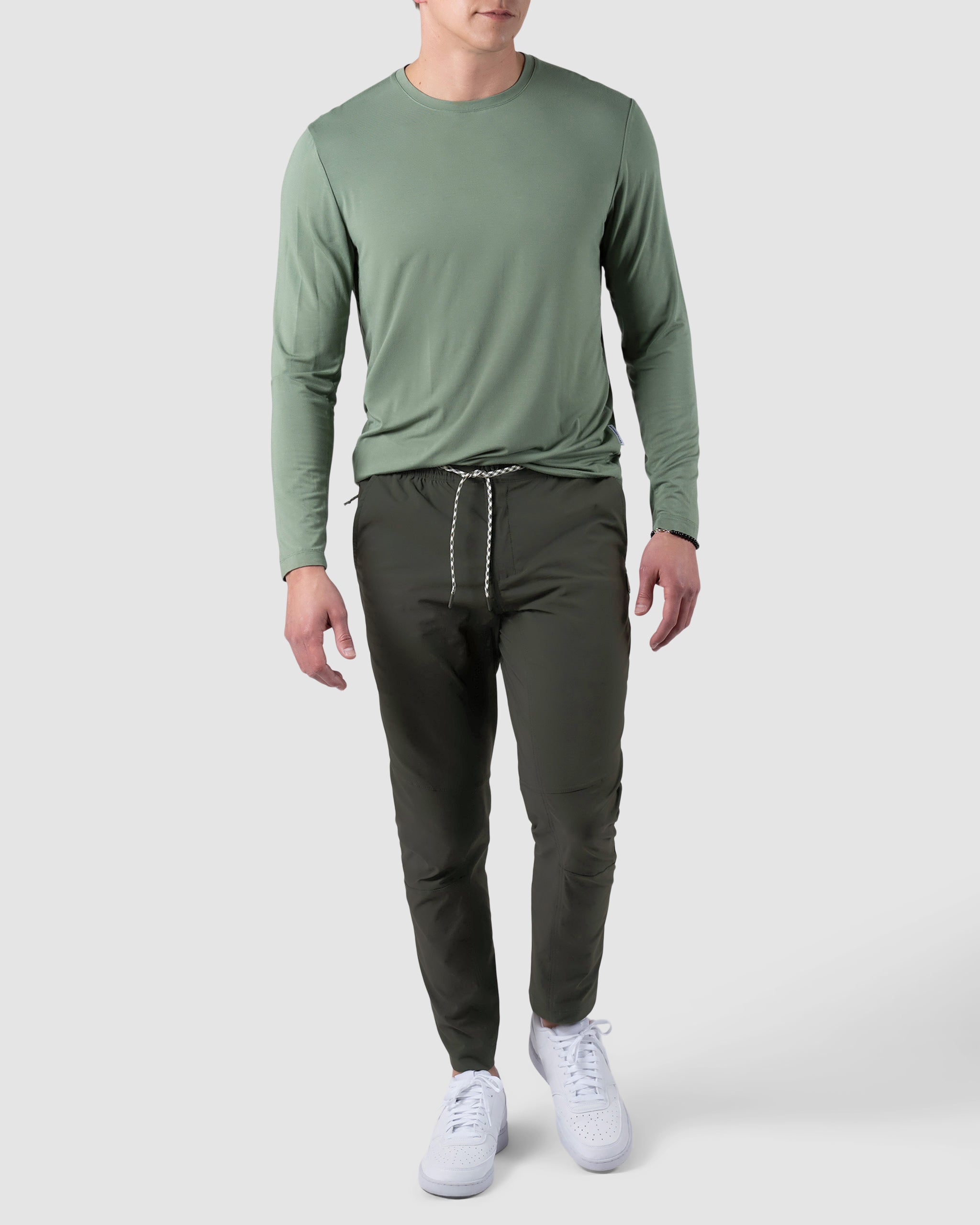A man wears an olive green long-sleeve shirt, BEARDEDGOAT NEW—ROVER™ Pant in dark green with a drawstring, and white sneakers, walking against a light gray background. His head is not fully visible.