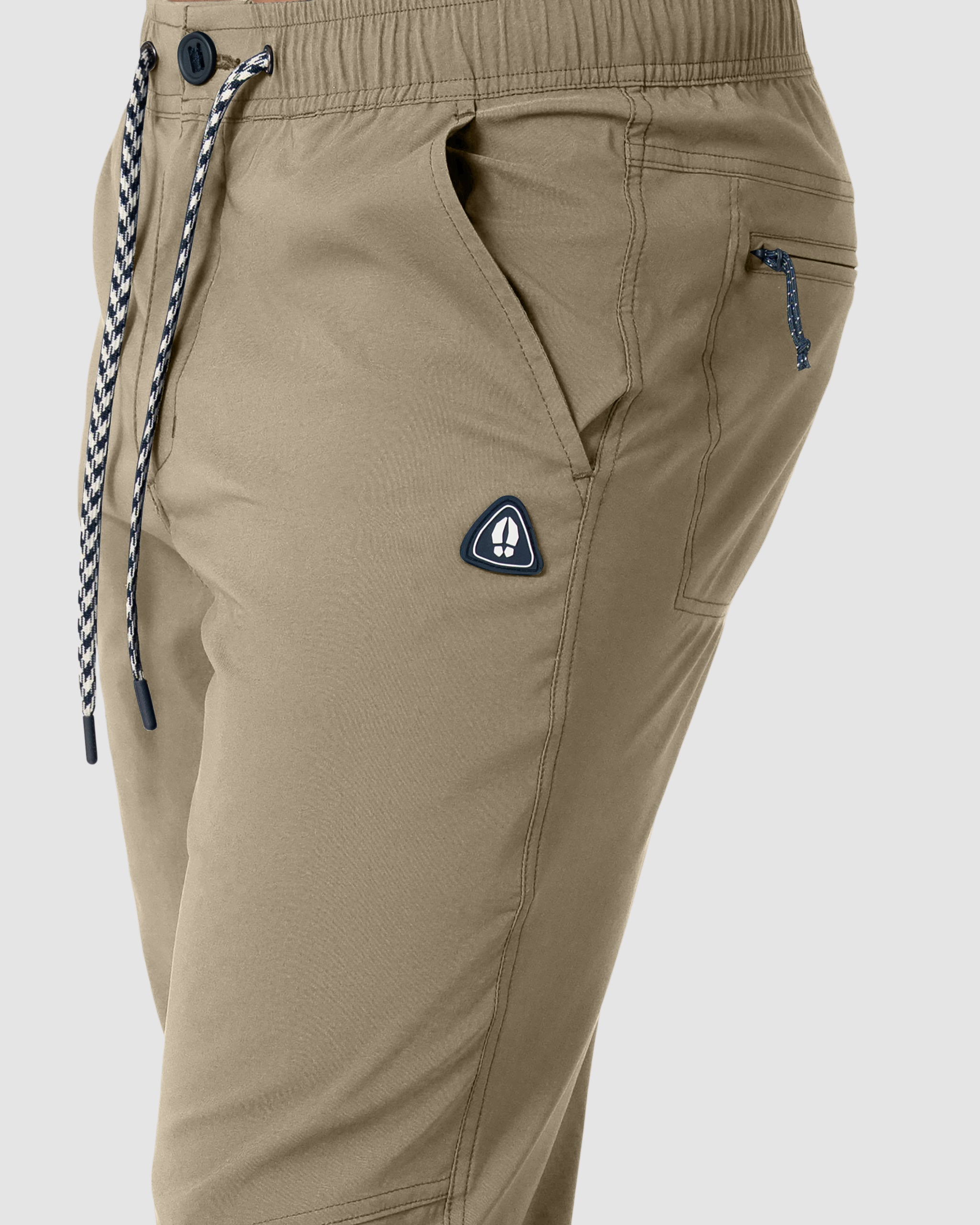 Close-up of a person wearing BEARDEDGOAT's NEW-—ROVER™ Pant in tan, made from performance stretch nylon with black and white drawstrings, a zippered pocket, and a triangular logo patch—perfect durable outdoor joggers for any adventure.
