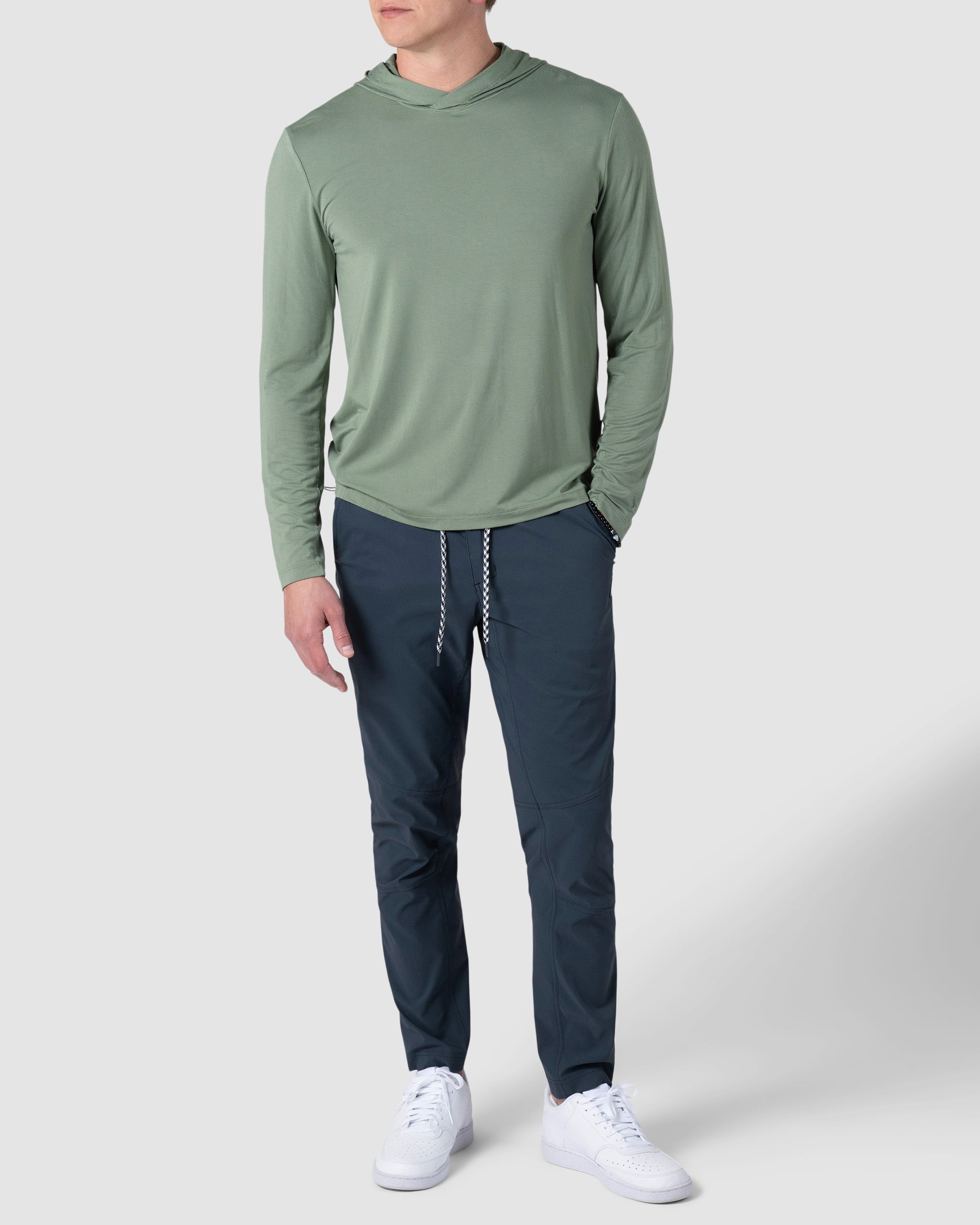 A man wearing BEARDEDGOATs Mens Rover Pant in dark navy, a light green long-sleeve hooded shirt, and white sneakers stands against a plain light gray background.