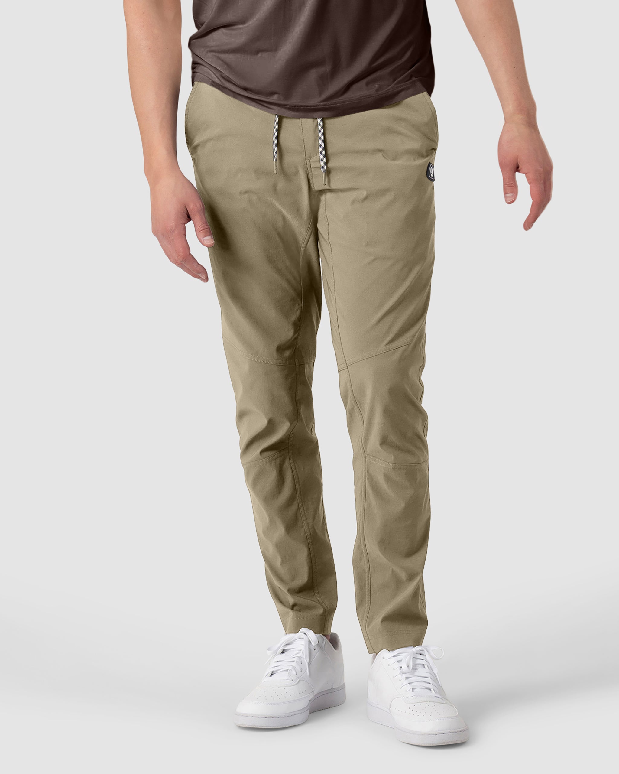 A person wears the BEARDEDGOAT NEW-—ROVER™ Pant in tan, styled jogger-style with a drawstring waist, paired with a brown shirt and white sneakers, standing against a plain light background. The head is not visible in the image.