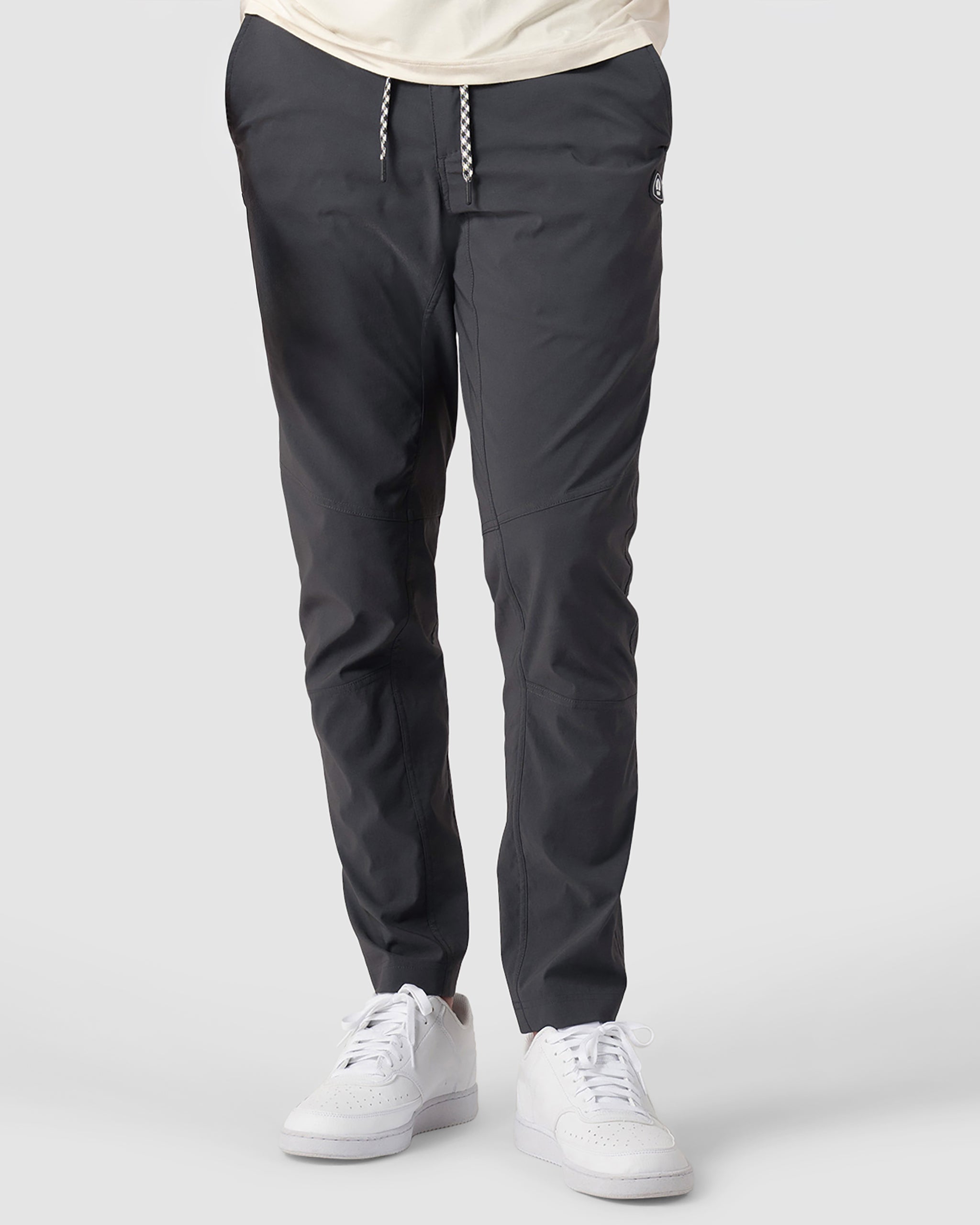 A person wearing BEARDEDGOAT NEW-—ROVER™ Pant in dark gray with a white drawstring and white sneakers, standing against a plain light gray background. Only the lower half of their body is visible.