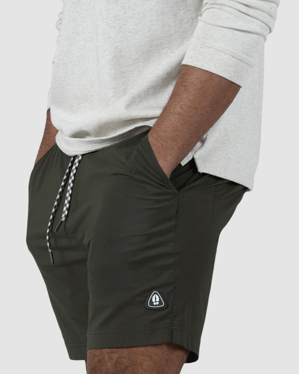 A person stands sideways with hands in pockets, wearing a light long-sleeve shirt and BEARDEDGOAT Mens Rover Hybrid Short in dark green. The stretch nylon shorts have a drawstring, small logo patch near the hem, and a plain light gray background.