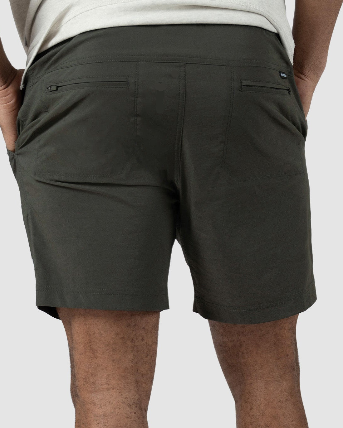 A person stands with hands in pockets, seen from behind against a plain light background, wearing BEARDEDGOAT Mens Rover Hybrid Short in dark green, paired with a light shirt, white socks, and white sneakers.
