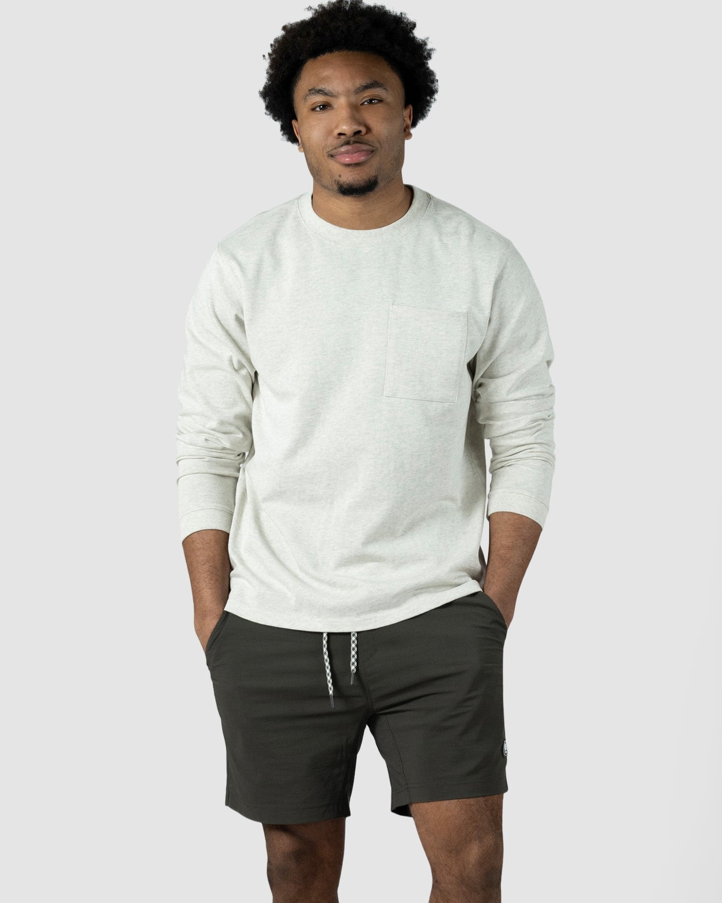 A man stands against a plain light background wearing a light beige long-sleeve shirt and BEARDEDGOAT Mens Rover Hybrid Shorts in dark olive green stretch nylon with a drawstring, paired with white sneakers. His hands are in his pockets and he has a neutral expression.