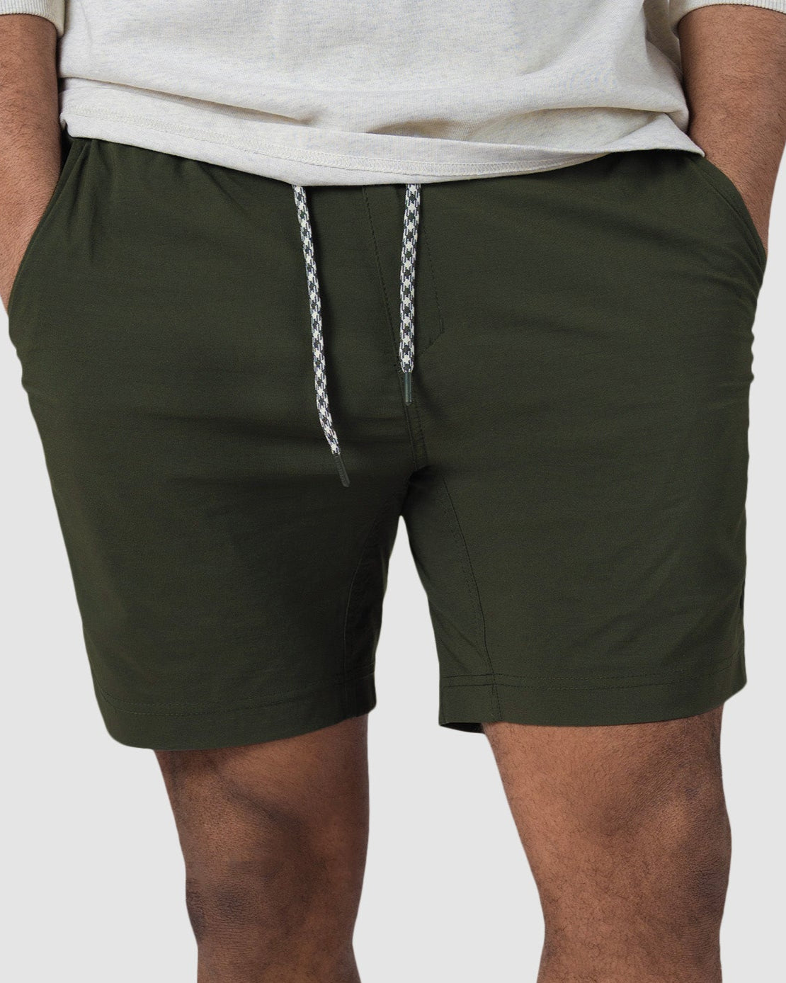 A person stands against a plain light background wearing BEARDEDGOAT Mens Rover Hybrid Shorts in olive green stretch nylon, paired with a light shirt, white socks, and white sneakers, hands in pockets.
