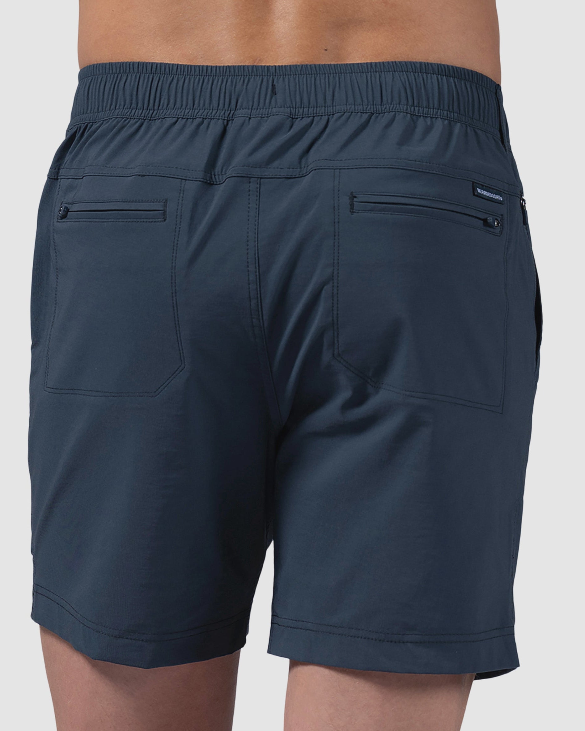 A man models the BEARDEDGOAT Mens Rover Hybrid Short in dark blue stretch nylon, with an elastic waistband, back pockets, and zippered compartments, photographed from behind against a plain light background.