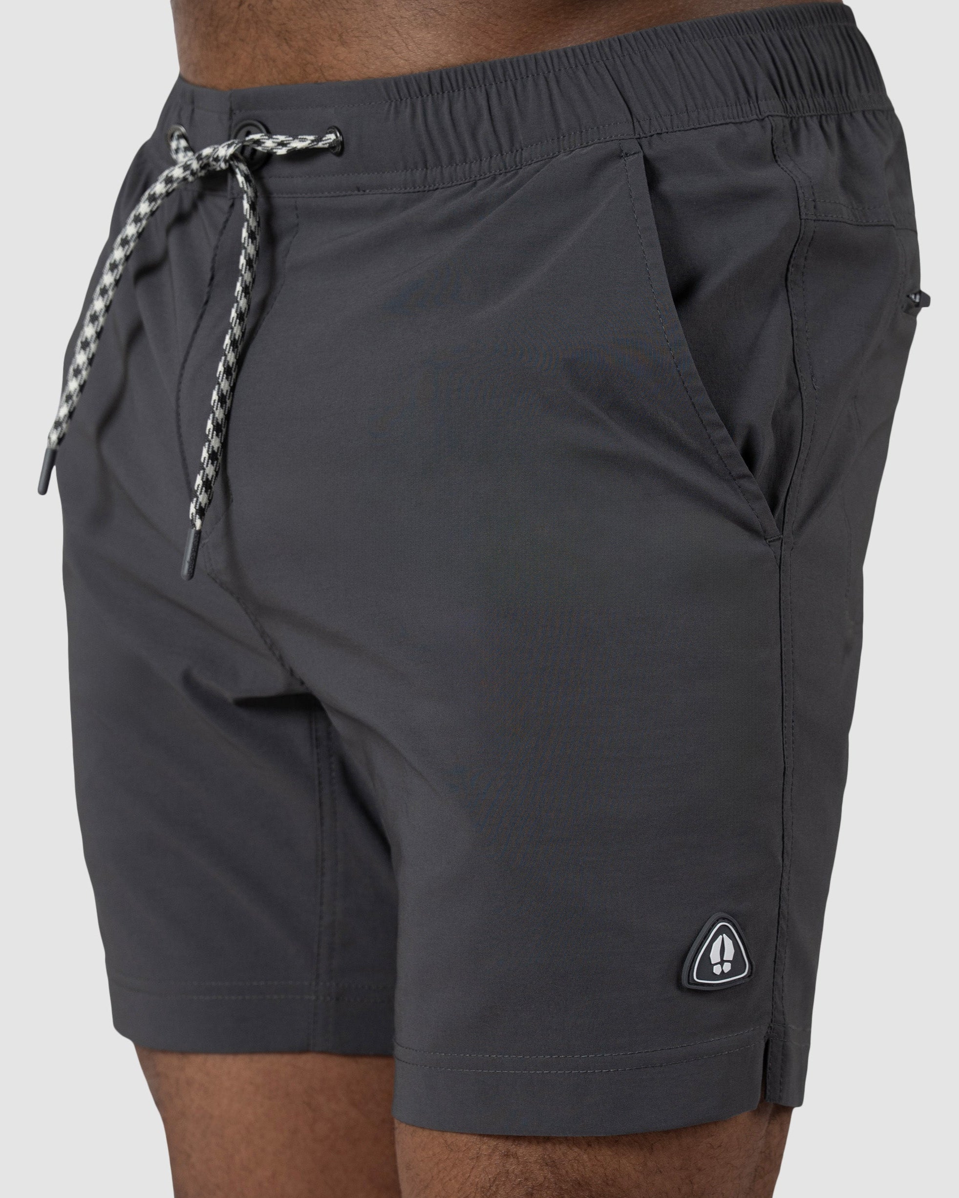 A person wears BEARDEDGOAT Mens Rover Hybrid Short—black stretch nylon with hands in pockets. These high-performance shorts feature a small triangular logo patch near the hem and white-and-black patterned drawstrings.