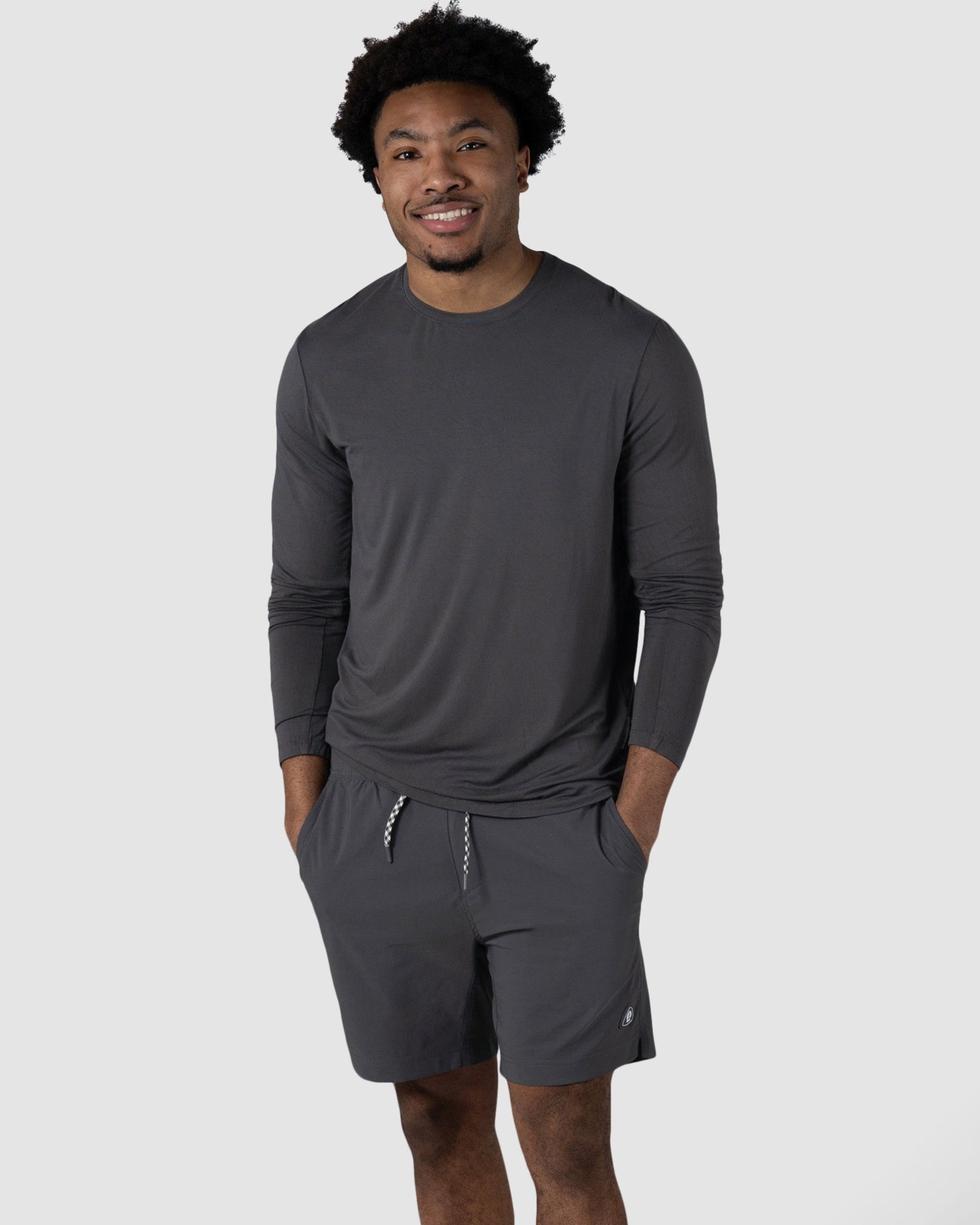 A man faces forward, smiling, wearing a long-sleeve dark gray shirt and BEARDEDGOAT Mens Rover Hybrid Short made from high performance stretch nylon, paired with white sneakers, against a plain light gray background.