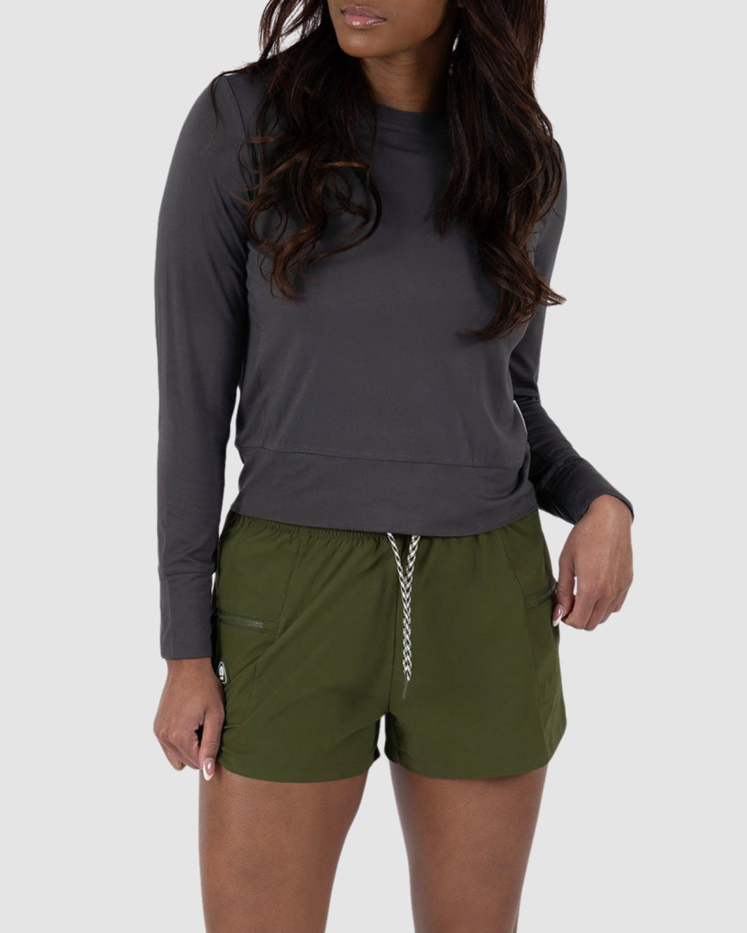 A woman with long, wavy hair stands against a light gray background, wearing the BEARDEDGOAT Womens Summit Short in olive green and a dark gray long-sleeve shirt. The stretchy ripstop shorts are paired with white sneakers for a relaxed look.