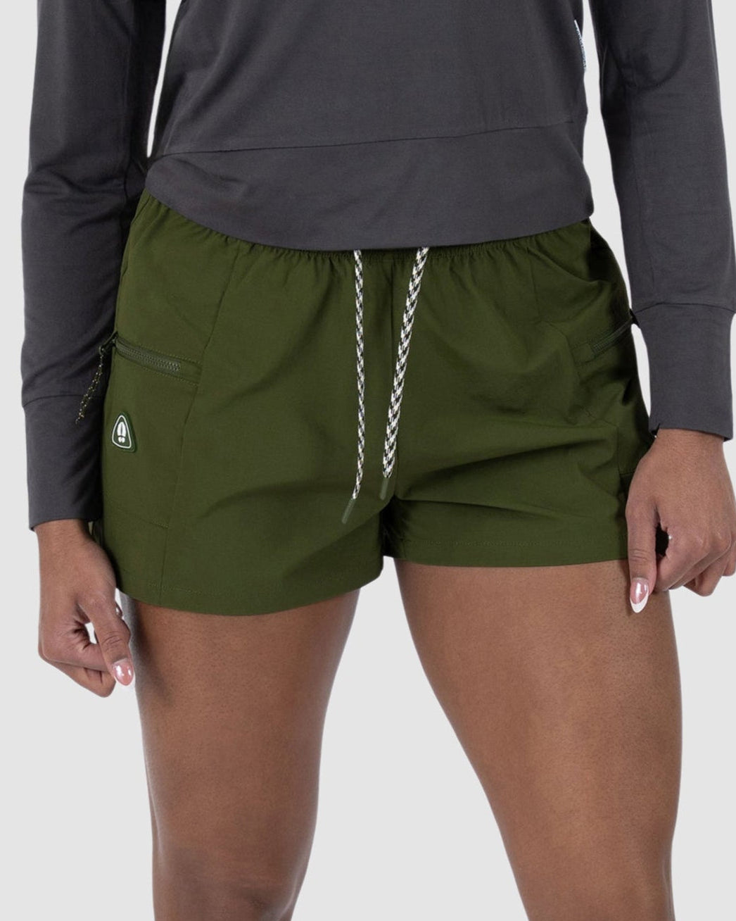 A person, visible from mid-torso down, wears a long-sleeve dark gray shirt and BEARDEDGOAT Womens Summit Short in olive green—made from lightweight stretchy ripstop fabric—plus white sneakers, standing against a light plain background.