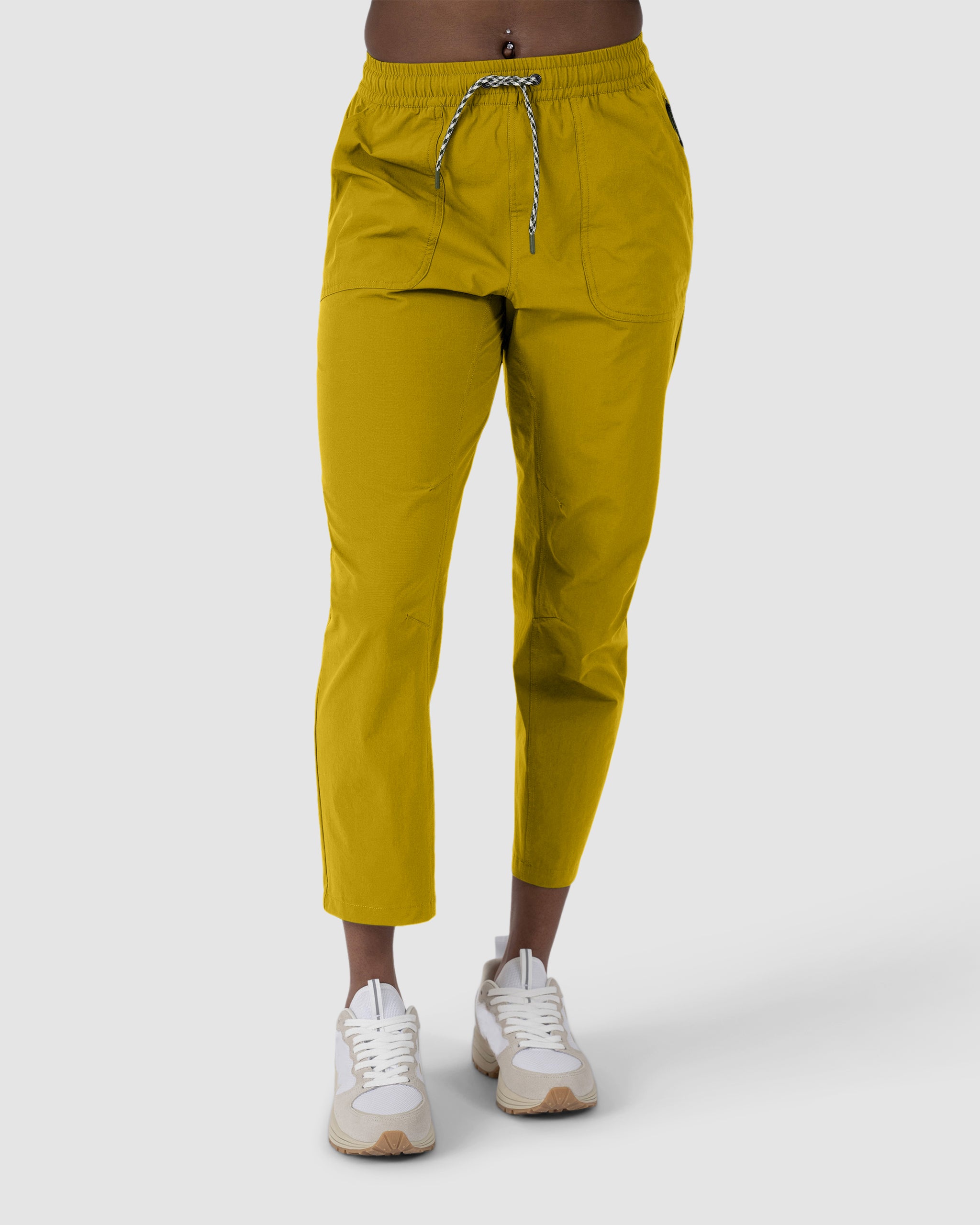 A woman in BEARDEDGOAT’s NEW — Summit™ Pant in Golden Palm, paired with white sneakers, stands against a plain light gray background.