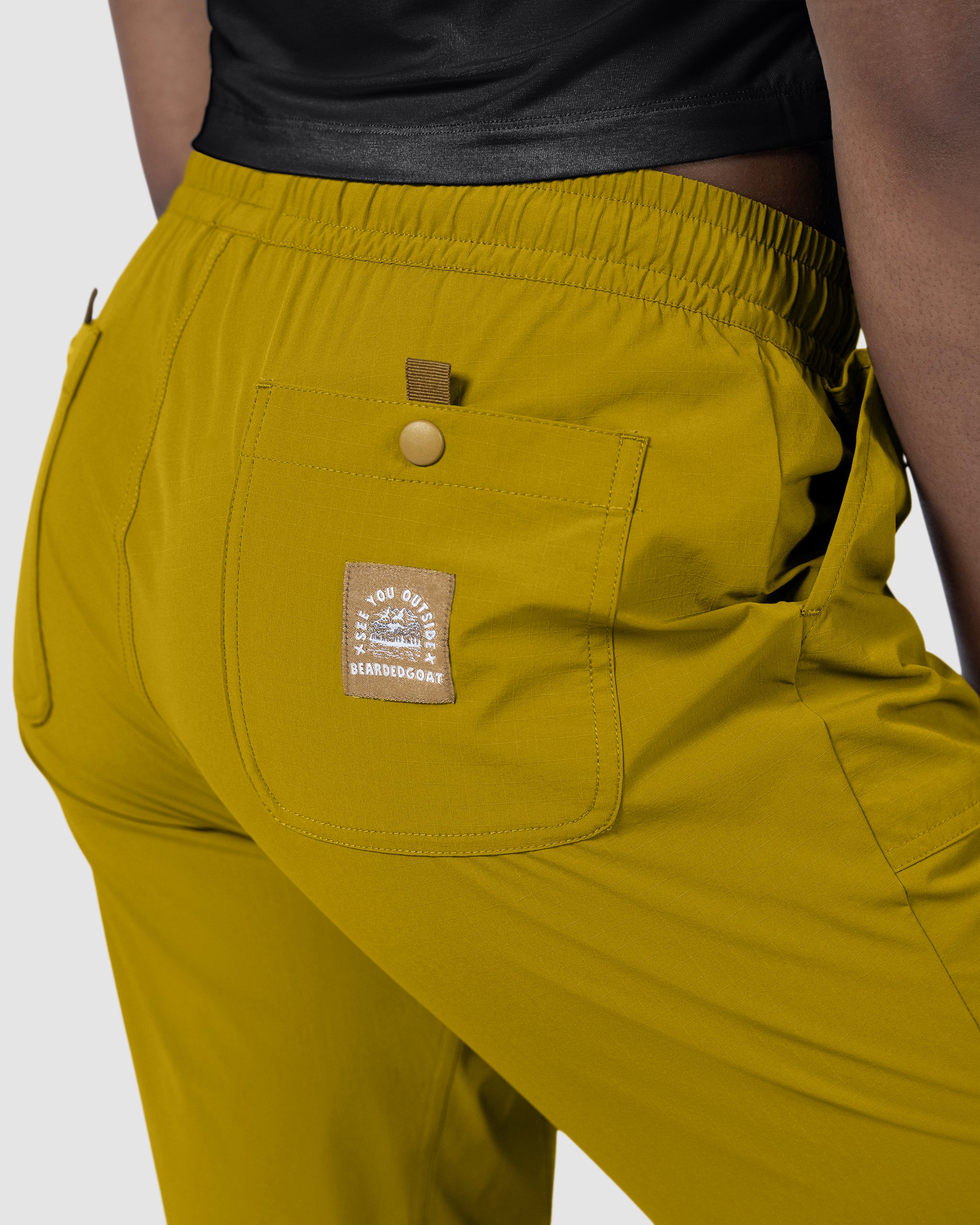 A close-up shows a person in mustard yellow BEARDEDGOAT NEW — Summit™ Pant, a women's performance style with an elastic waistband and back pocket patch, paired with a black top.