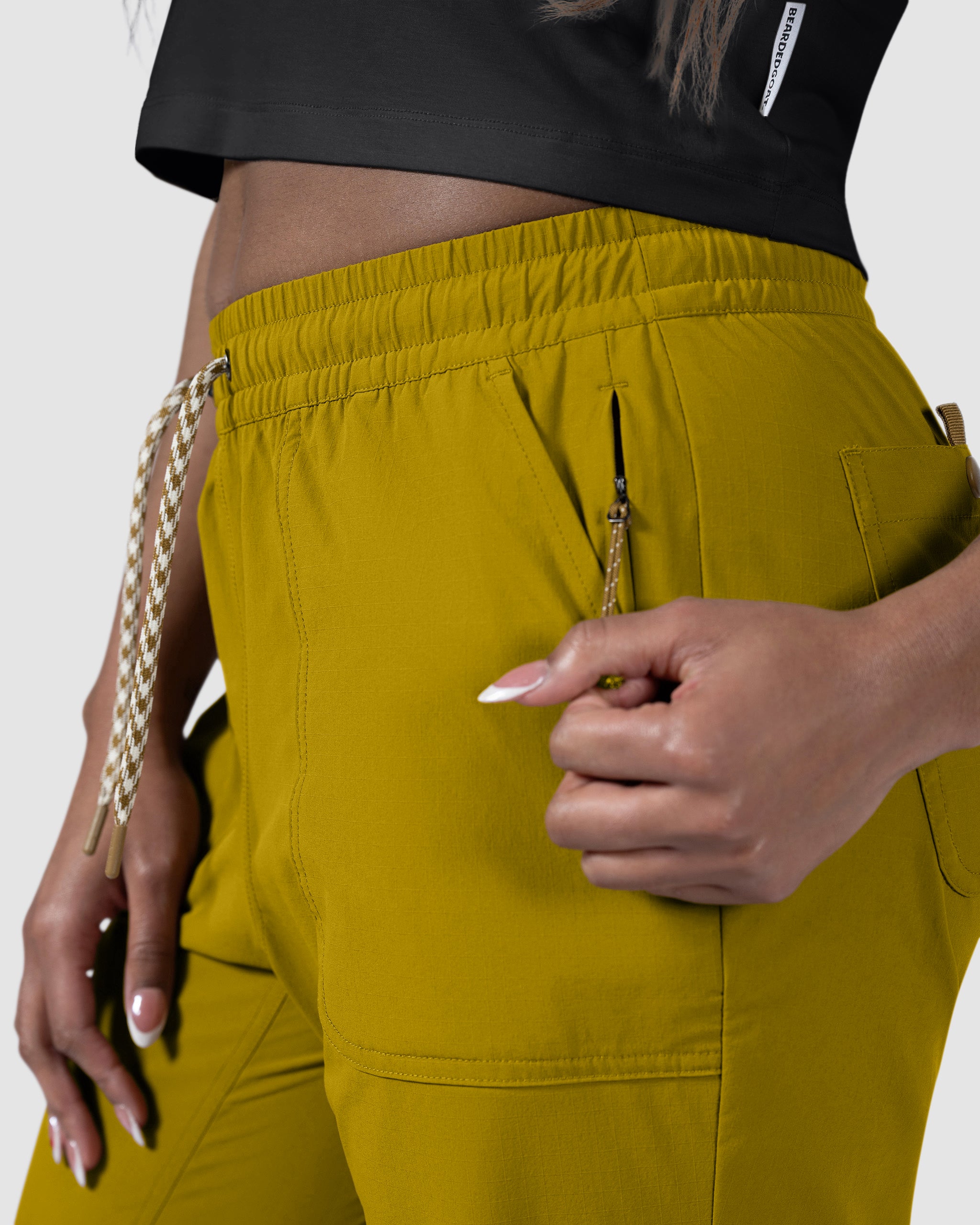 A person in BEARDEDGOAT NEW — Summit™ Pant in mustard yellow and a black crop top is unzipping a pocket on the women's performance pant. Only their torso and hands are shown.
