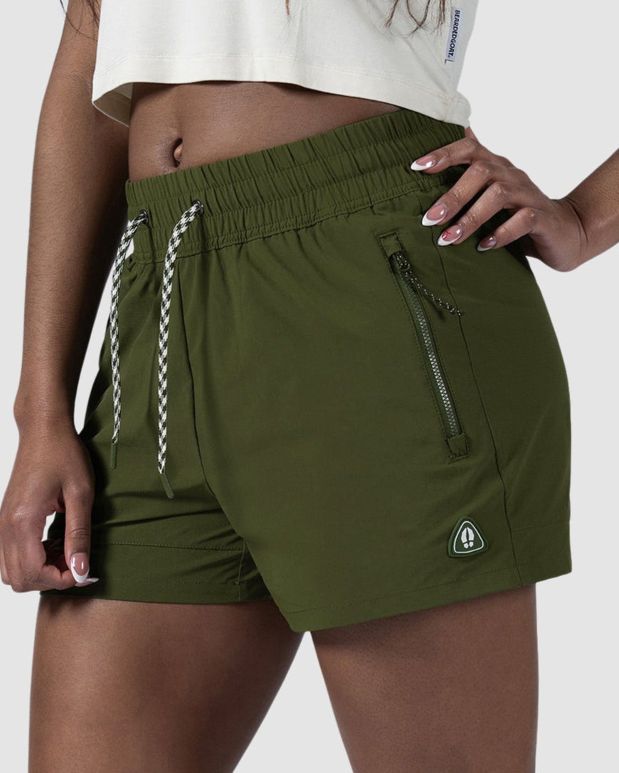 A woman stands with one hand on her hip against a plain light background, wearing a white crop top, BEARDEDGOAT Womens Alpina Short in olive green ripstop with zippered pockets, and white sneakers.