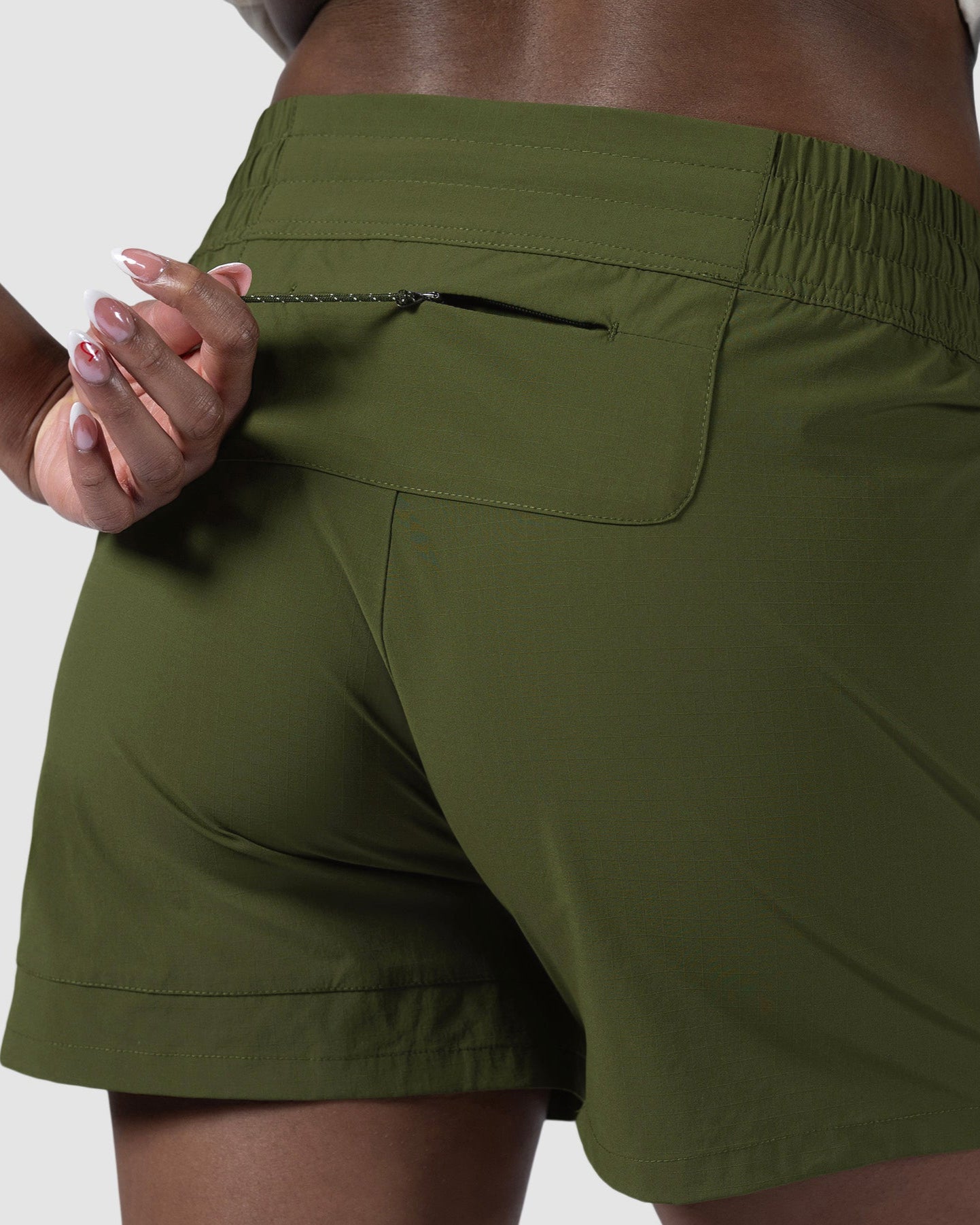 A person wearing BEARDEDGOAT Womens Alpina Short in olive green zips the back pocket. The image spotlights the high-waisted shorts zippered pockets from the waist down, set against a plain light background.