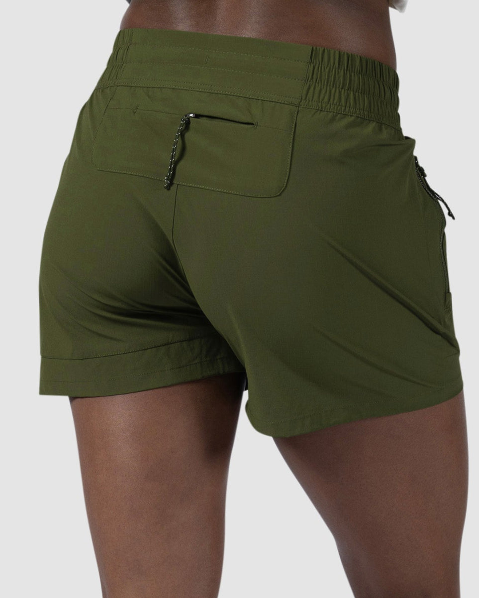 A person faces away, wearing BEARDEDGOAT Womens Alpina Short in olive green—high-waisted, stretchy ripstop fabric with zippered pockets—paired with white sneakers against a plain light gray background.