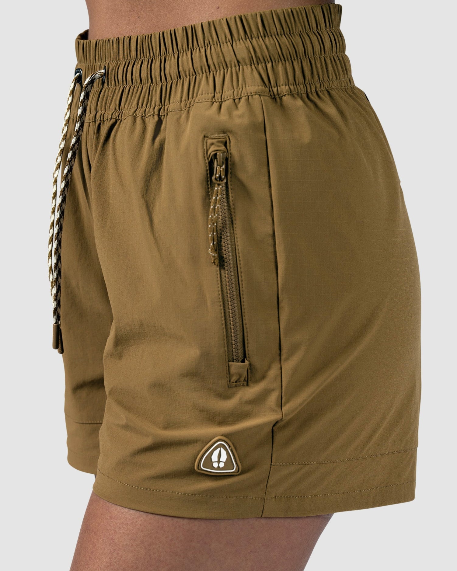 A woman shown from the side wears BEARDEDGOAT Womens Alpina Short—brown, high-waisted adventure shorts in stretchy ripstop fabric with a zippered pocket and a small triangular logo by the hem—against a plain background.