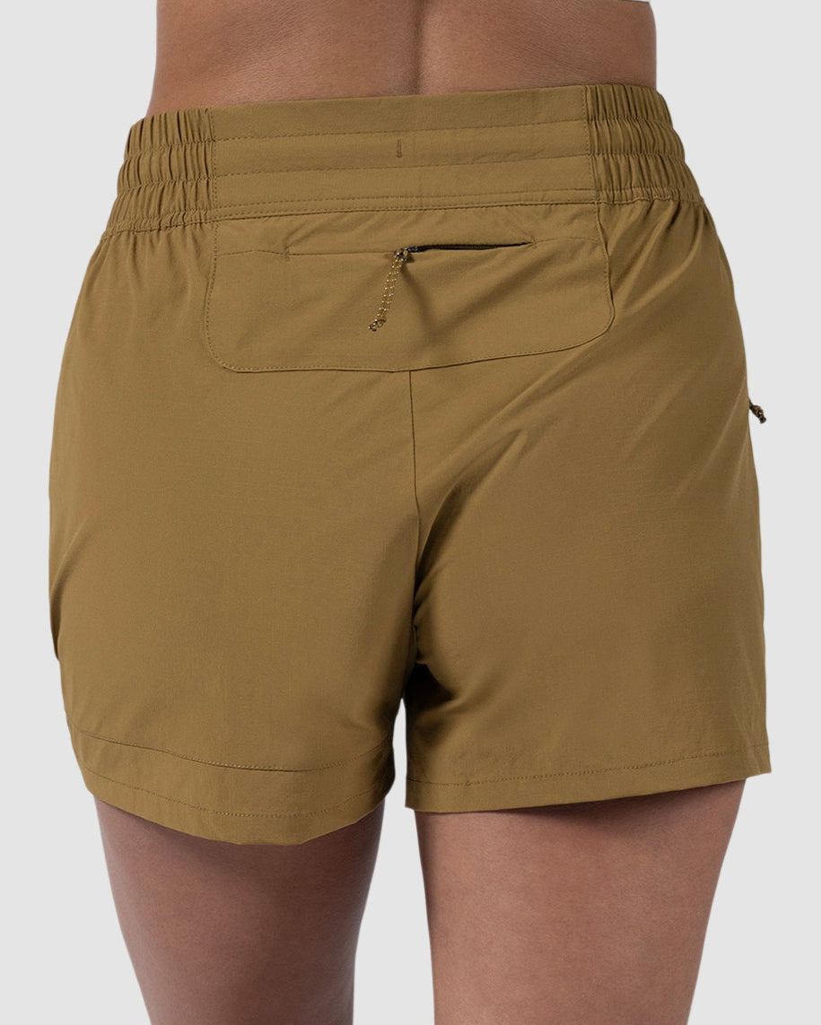 A person is shown from the back wearing BEARDEDGOAT Womens Alpina Short in olive green stretchy ripstop fabric and white sneakers, standing on a plain light background.