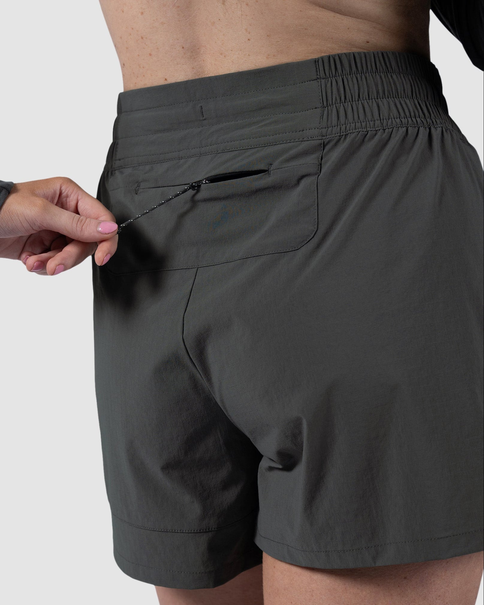 A person in BEARDEDGOAT Womens Alpina Short and a long-sleeve top unzips a pocket on the shorts, with only their lower back and hand visible against a plain gray background.